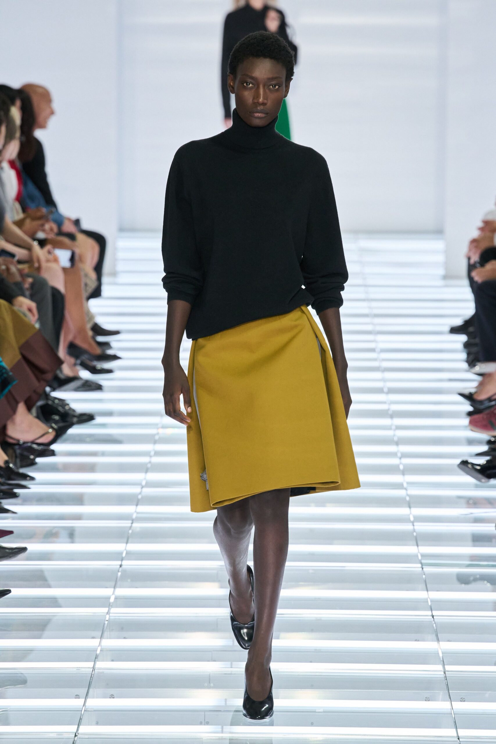 Black fine-knit turtleneck, sleeves pushed into soft bunching at the wrists. Mustard felted skirt cut in a stiff wrap A-line with folded under hem. Black patent pumps. Walking through a museum atrium before the heating has fully come on.