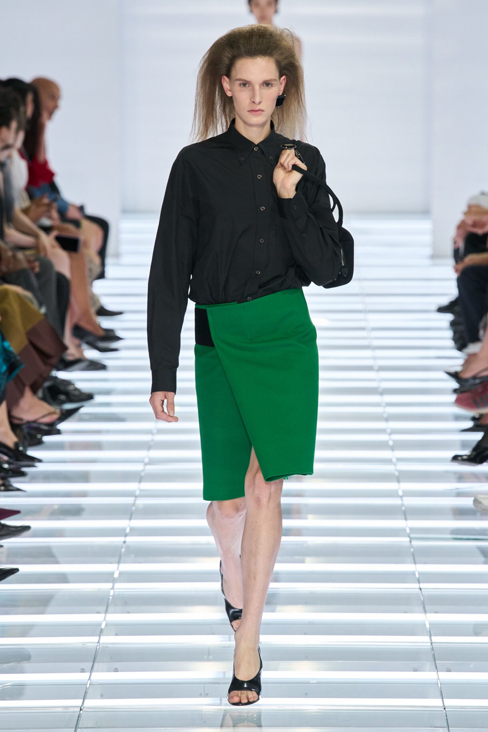 Black button-front shirt in crisp cotton, collar points pinned close. Kelly green wrap skirt with a sharp overlap and black side inset. Small black shoulder bag with a narrow strap tucked in hand. Black patent peep-toe mules. Walking into a municipal hearing with your notes still warm from the printer.