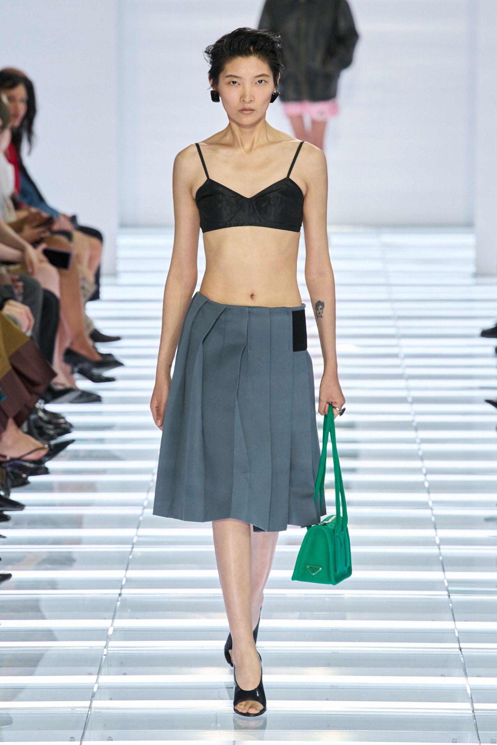 Chunky black square clips tight to the lobe. Black satin bralette with seamed cups and pin-thin straps. Blue-grey pleated skirt, one black inset panel breaking the waistline. Acid-green shoulder bag with a long doubled strap. Black peep-toe mules. Lunch break on a plaza so bright the paving throws light back up.