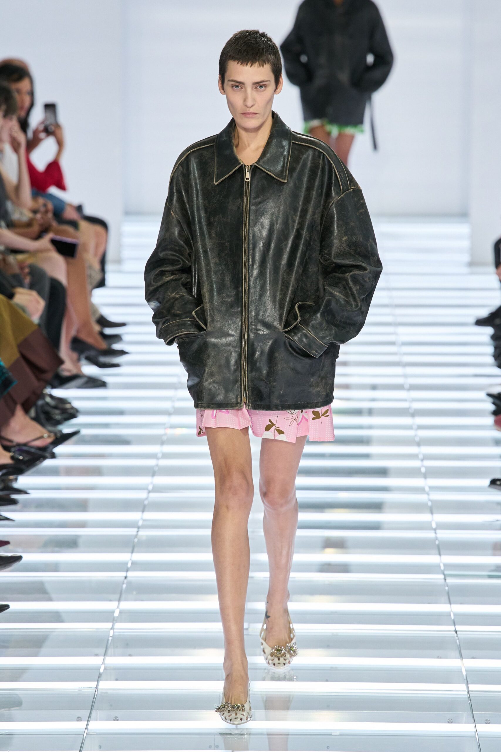 Oversized black leather jacket, rubbed finish and pale topstitching tracing the dropped shoulder. Pink printed mini peeking below, soft ruffle hem kicked uneven at the thigh. White cutout flats with jeweled clusters at the toe. Walking home from a motel wedding with the jacket borrowed off someone taller.