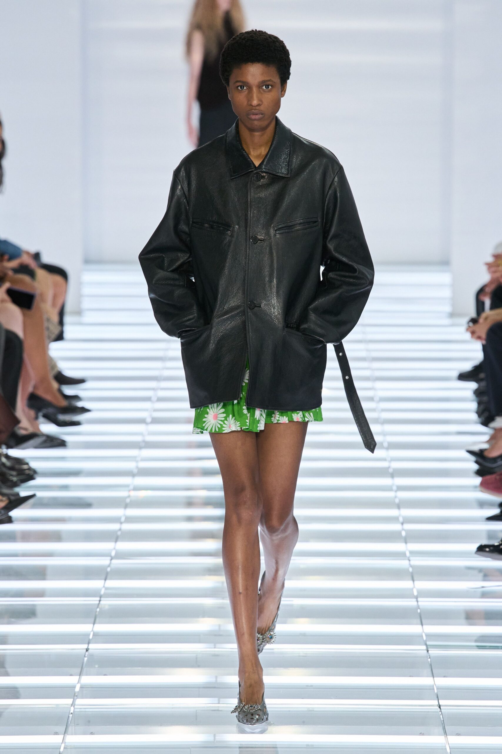 Oversized black leather jacket with welt chest pockets and a dangling side strap. Green daisy-print mini peeking below the hem in a soft ruffle. Silver mesh pumps topped with clustered crystal florets. Crossing a hotel atrium early, florist buckets still lined by the service lift.