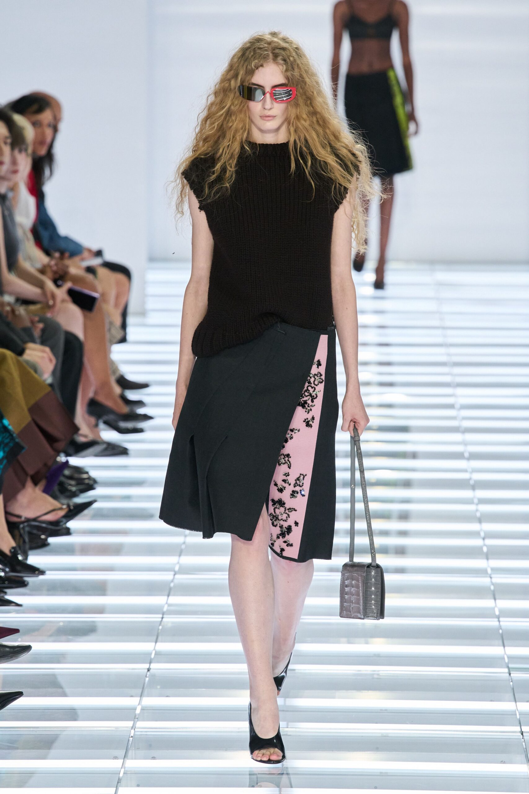 Angular wraparound sunglasses, one lens rimmed in hot pink. Chunky black knit sweater vest with raw cap sleeves. Asymmetric black skirt split by a pink sequined floral panel. Gray croc-embossed box bag on a thin strap. Black peep-toe mules. Crossing the hotel atrium before the wedding brunch turns noisy.