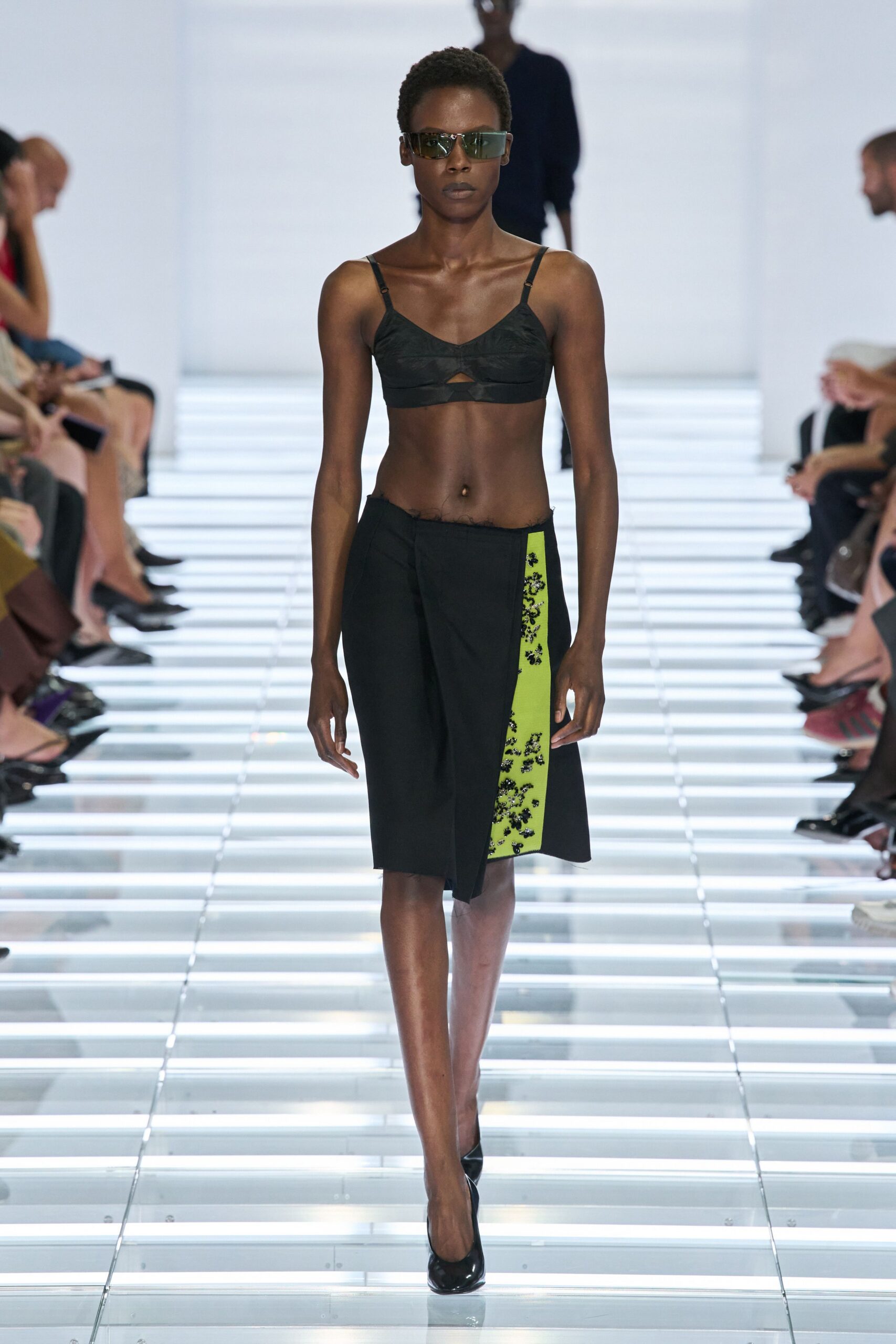 Wraparound visor shades with a smoky mirrored lens. Black bralette with gathered cups and a tiny underbust keyhole. Asymmetric black wrap skirt, chartreuse inset crusted with black beading. Black patent pumps. Crossing a glass concourse before the building opens for the day.