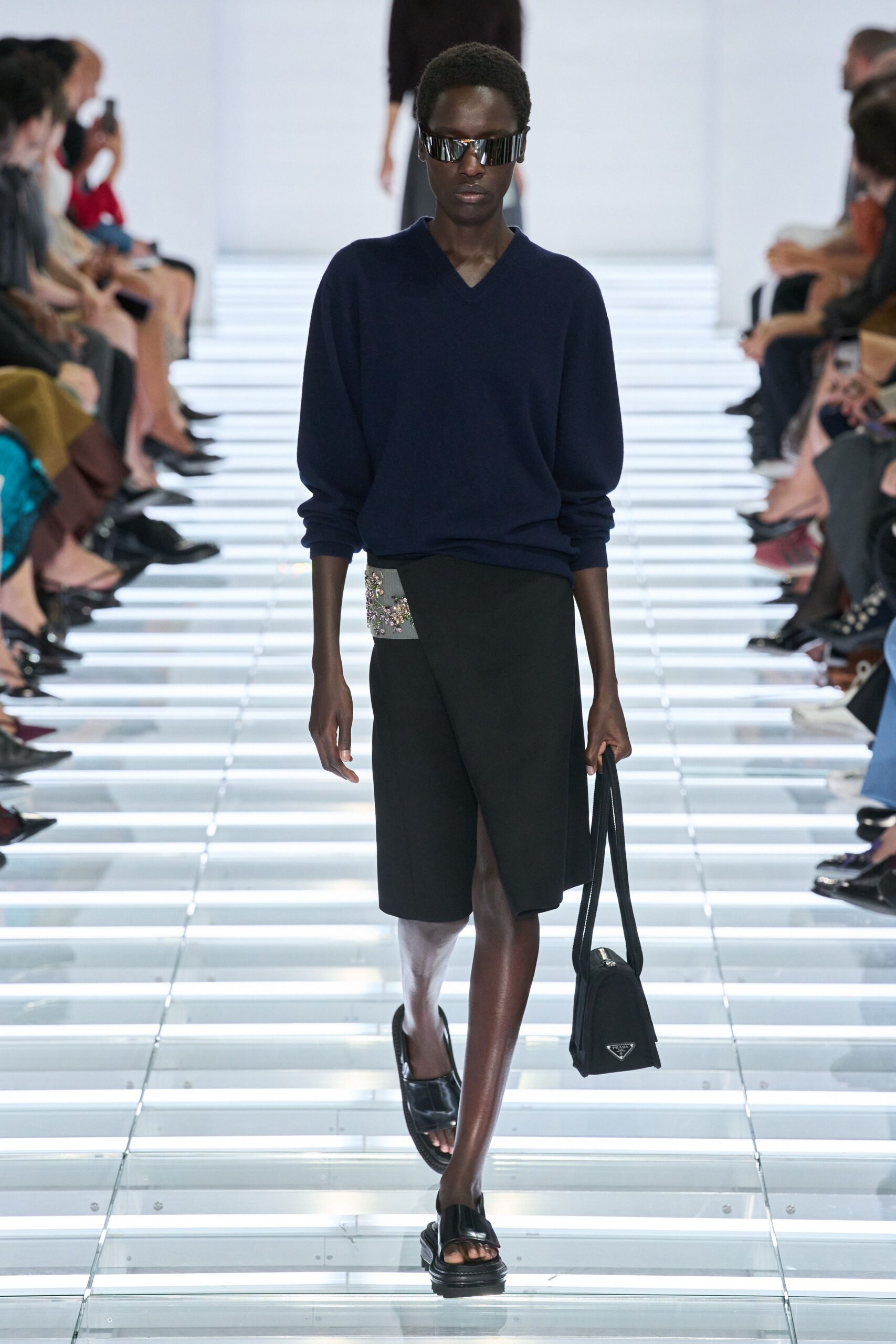 Wraparound visor shades with mirrored black slats. Navy fine-knit V-neck sweater, hem slouching slightly off center. Black wrap skirt with a grey beaded hip panel. Long-strap black nylon bag, narrow and boxy. Black leather slide sandals with thick molded soles. Crossing a glass footbridge before the office towers fully wake.
