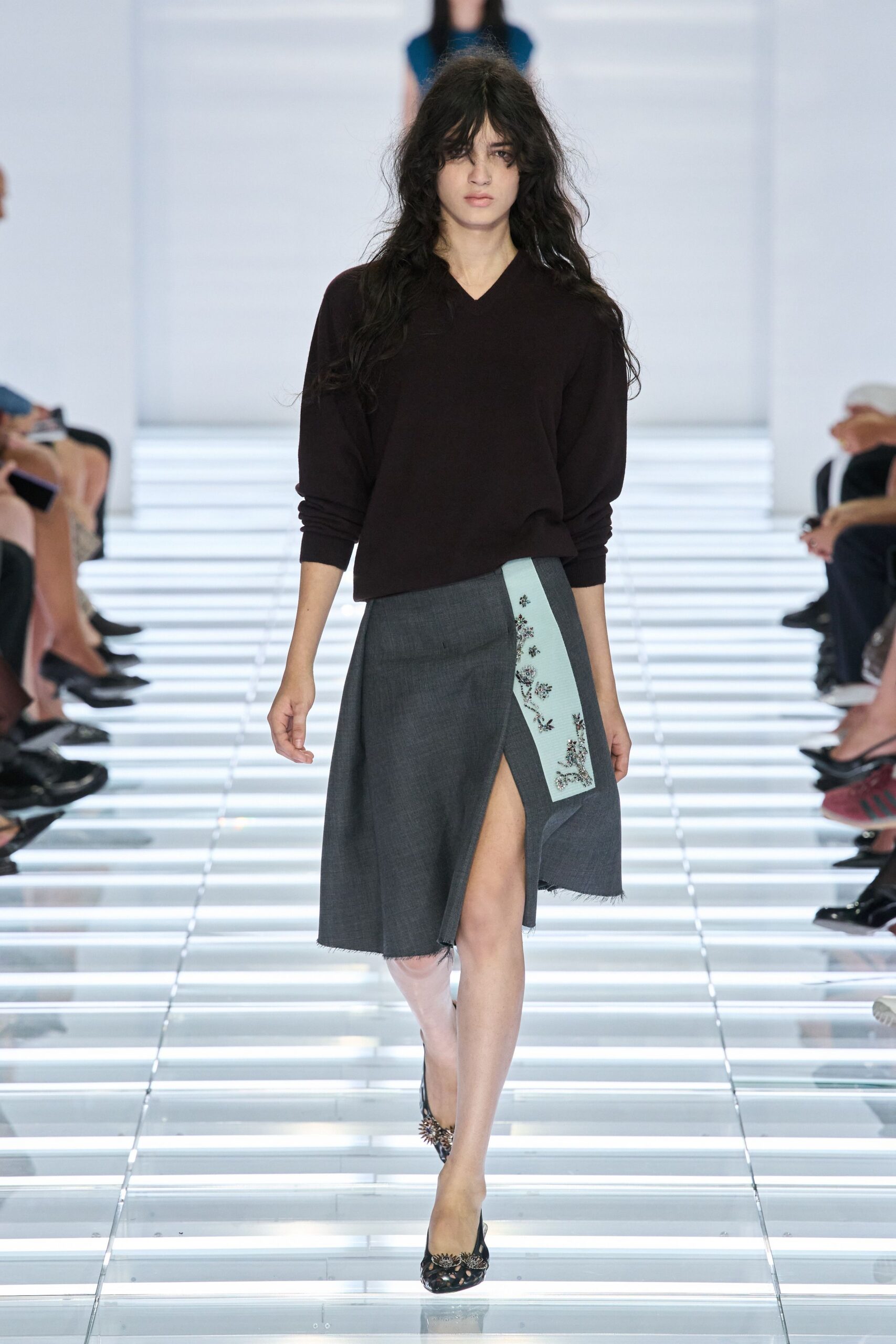 Dark espresso knit sweater with a narrow V and pushed sleeves. Charcoal wrap skirt in suiting wool, frayed hem and thigh-high overlap. Pale blue grosgrain panel beaded with small floral sprays. Black cutout heels tipped with clustered crystals. Crossing a glass atrium before the museum opens to the public.