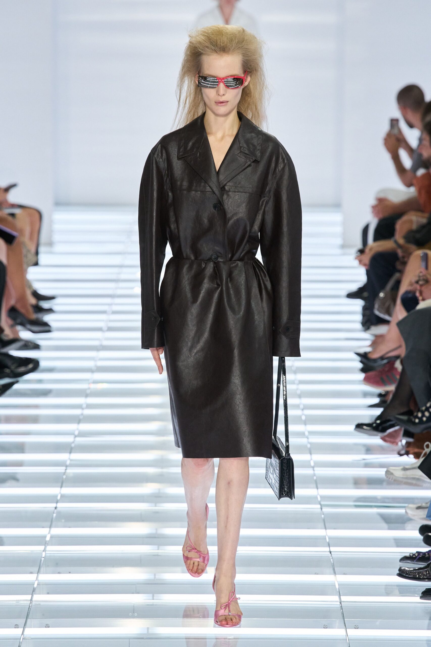 Red wraparound visor shades with white slatted lenses. Black leather coat dress, broad lapels and a pinched one-button waist. Slim black crocodile-embossed shoulder bag hanging low at the hip. Pink thong sandals tied in small bows over the toes. Crossing a glass skybridge with office flowers still in your hand.