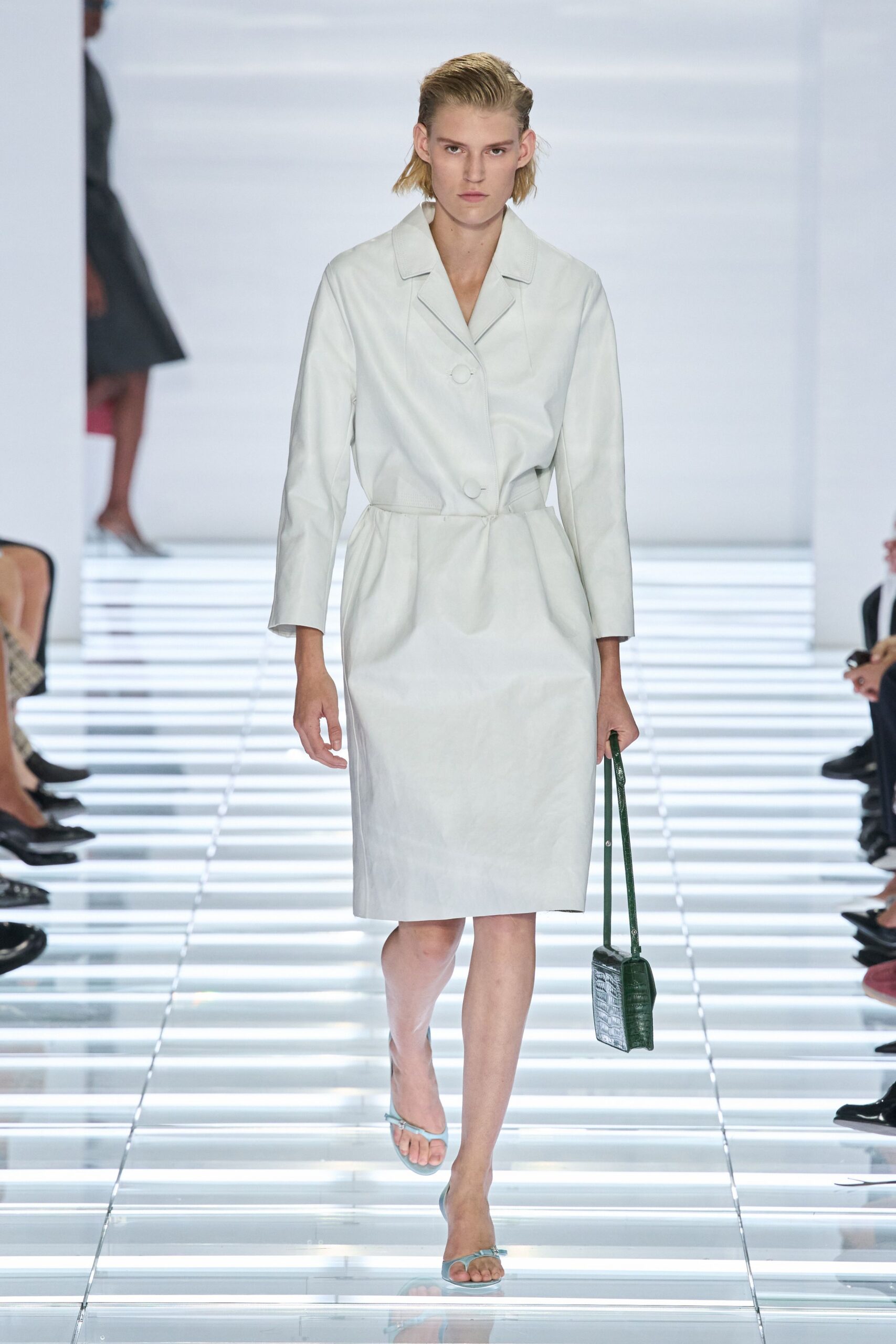 Pale celadon leather coatdress with a dropped waist and oversized covered buttons. Dark green crocodile-embossed box bag on a narrow shoulder strap. Icy blue thong sandals with clear lucite heels. Crossing a hotel atrium before breakfast, carrying only a room key.