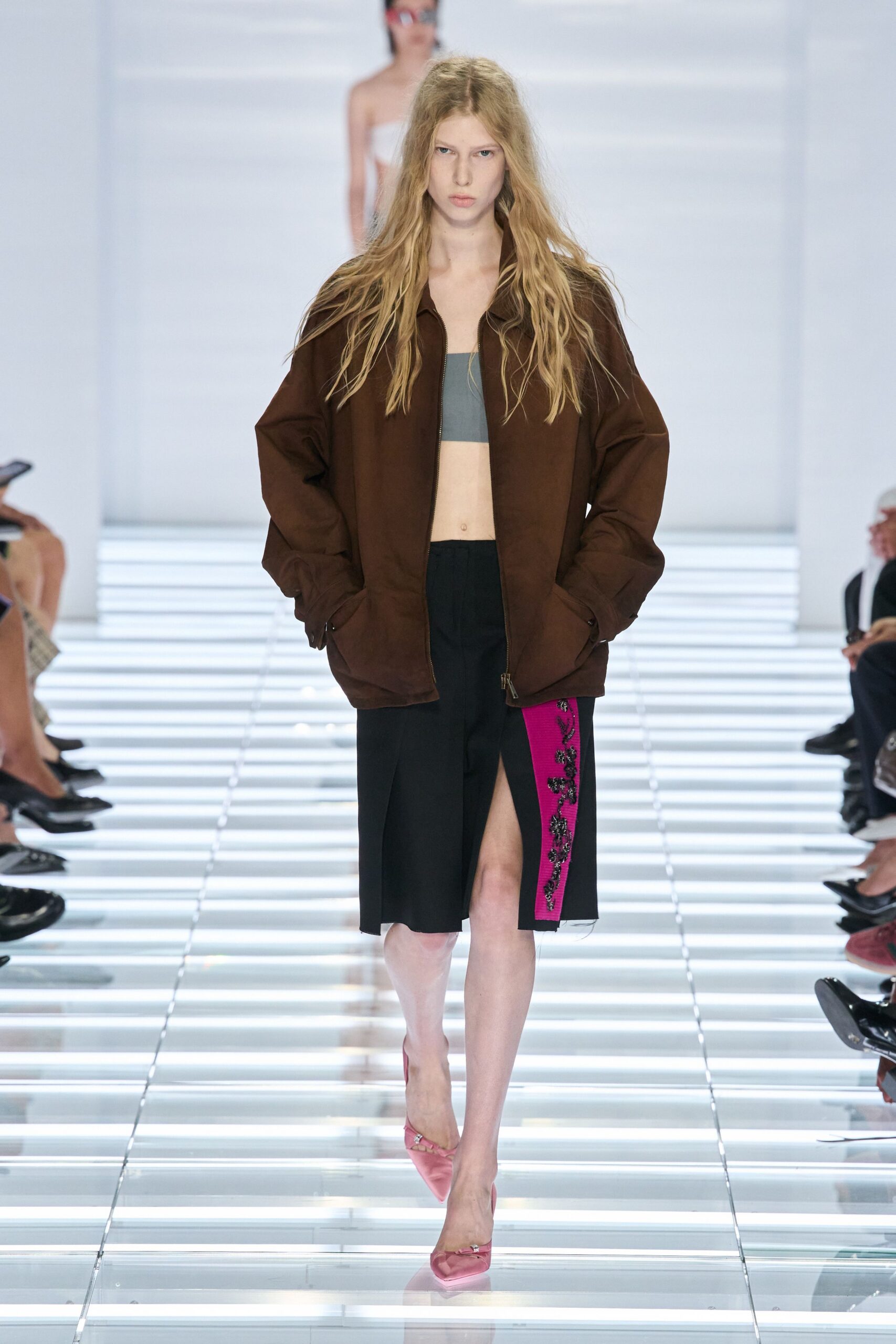 Oversized chocolate zip jacket with soft folds bunching at the cuffs. Slate bandeau cut straight across the chest. Black knee-length skirt split high, with a hot-pink beaded side panel. Pink satin pointed pumps with a tiny crystal buckle. Walking into a hotel lobby still carrying pool heat on your skin.
