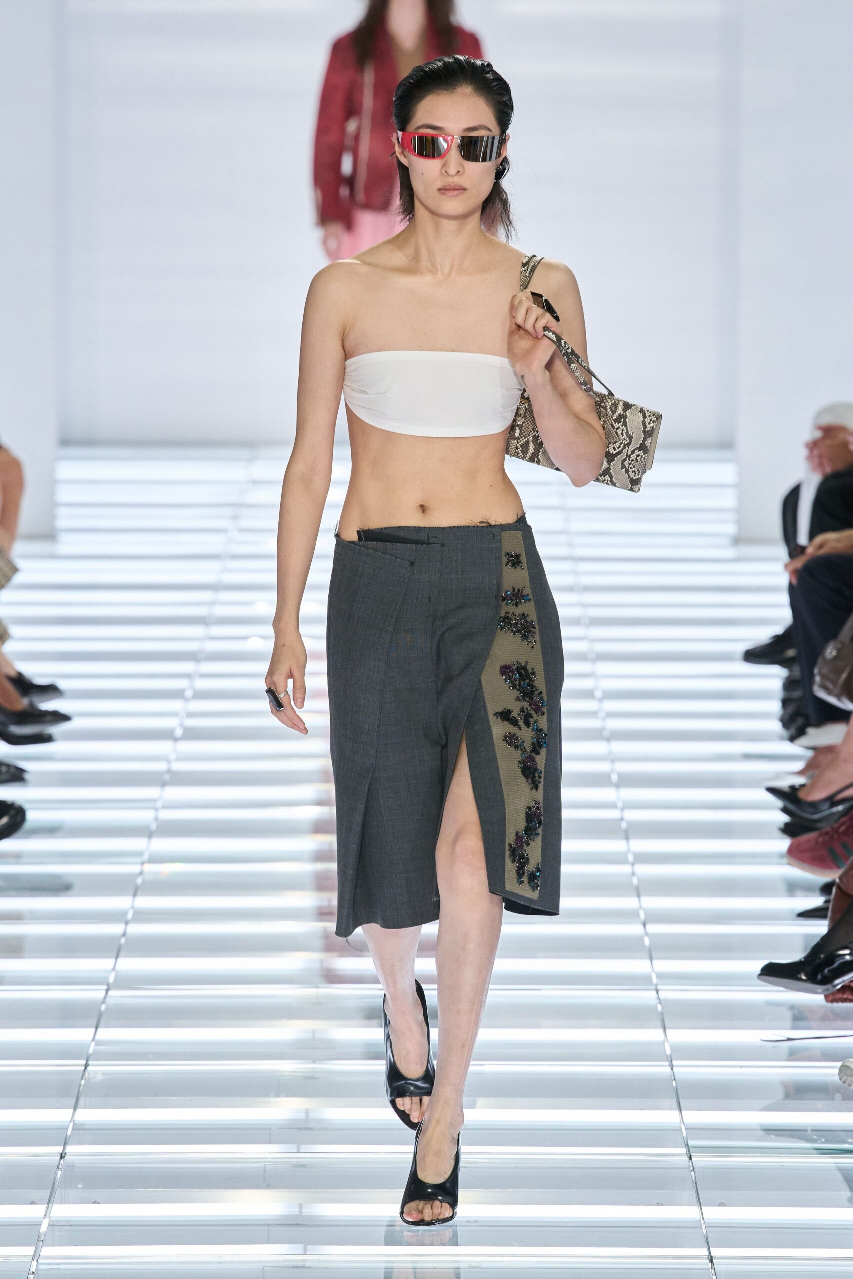 Wraparound mirror visor with a hot-pink brow bar. White stretch bandeau, ruched slightly at the side seams. Python-pattern shoulder bag tucked tight under one arm. Charcoal wrap skirt with raw waistband and beaded mesh side panel. Black peep-toe mules. Walking into a hotel lobby still carrying sand in your shoes.