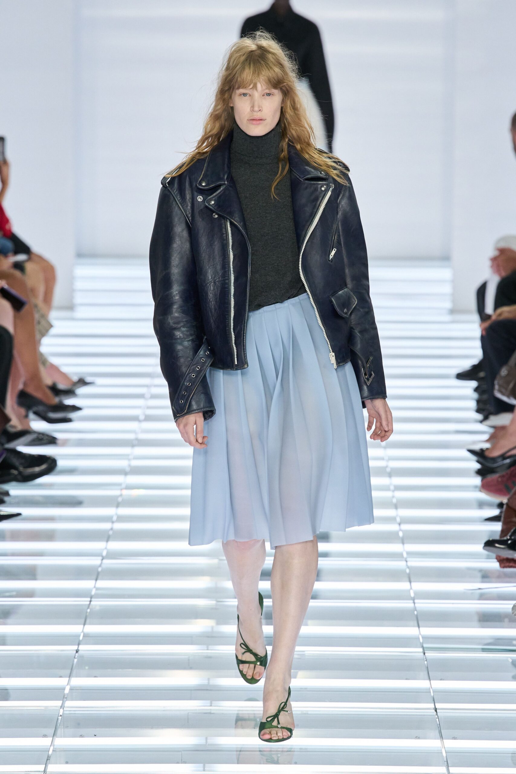 Ink-navy leather biker jacket with bright silver zip teeth and snapped lapels. Charcoal fine-knit turtleneck, close at the throat. Powder-blue pleated skirt falling in soft knife folds below the knee. Moss green suede sandals with bow-tied front straps. Crossing a glass atrium before the offices fill for Monday.