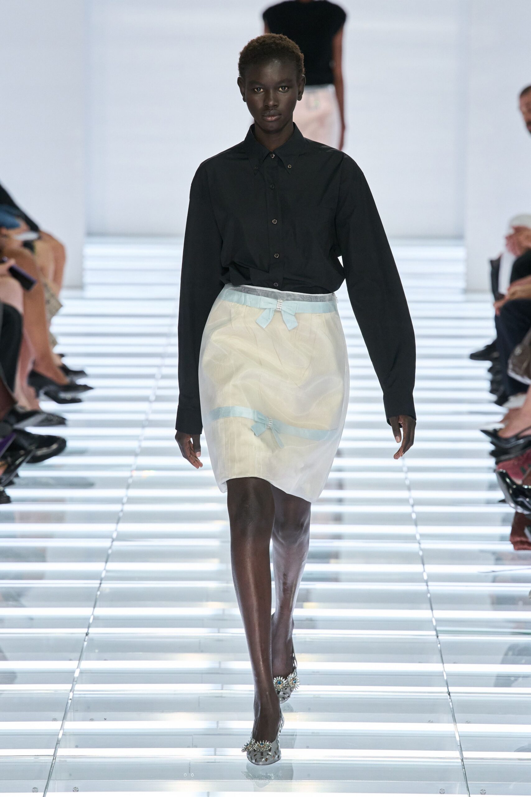 Black cotton button-down, collar tips pinned with tiny silver studs. Sheer ivory organza pencil skirt over a pale slip, banded with blue bows. Clear pumps topped with crystal rosettes at the toe. Crossing a hotel atrium early, before the florist has finished the urns.