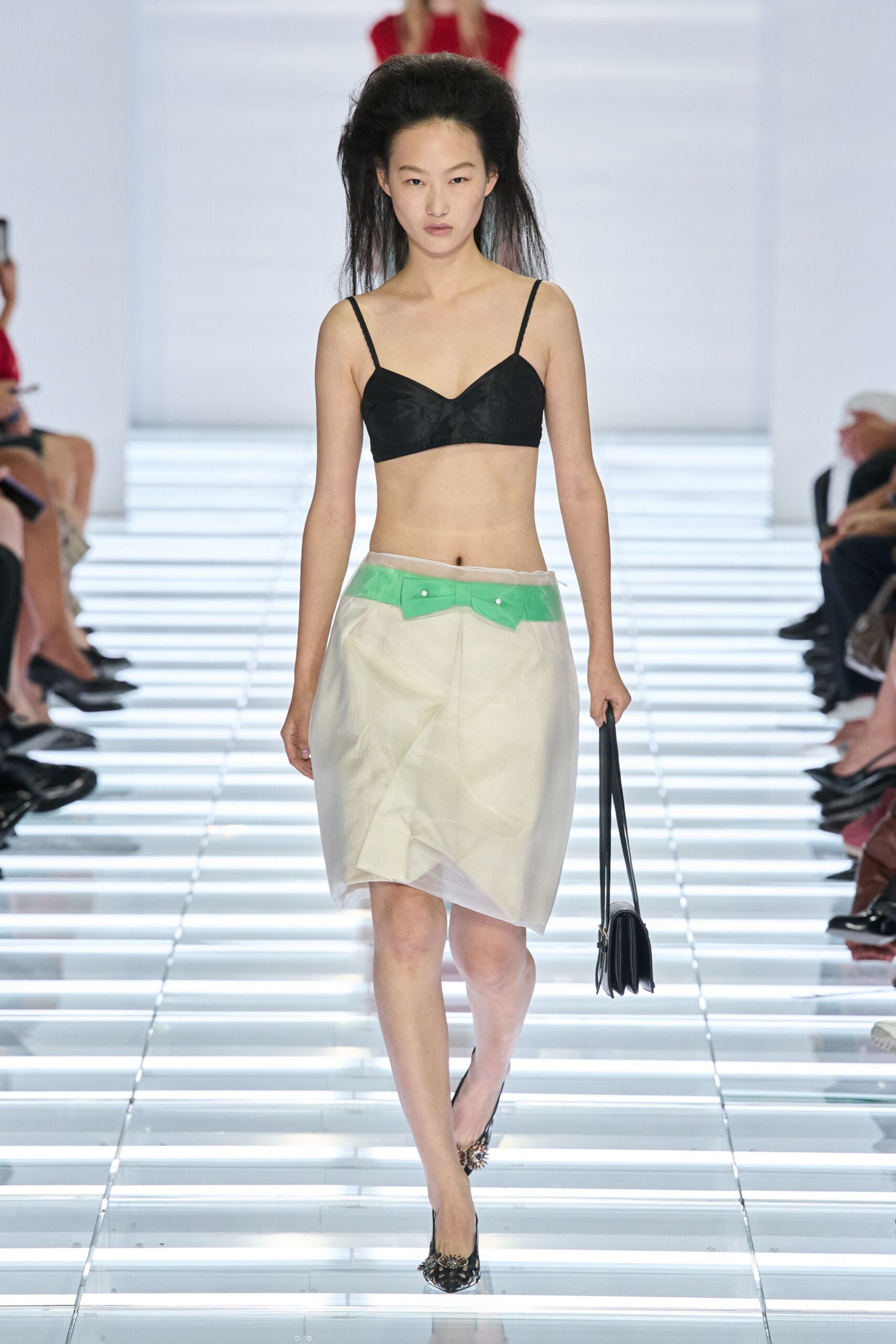 Black bra top with lightly mottled cups and narrow straps. Milky organza skirt over a cream slip, acid-green waistband tabs snapped on. Slim black bag hanging long and flat at the side. Pointed leopard pumps with a spiky cluster at each toe. Crossing a hotel atrium before breakfast, carrying only room key and nerve.