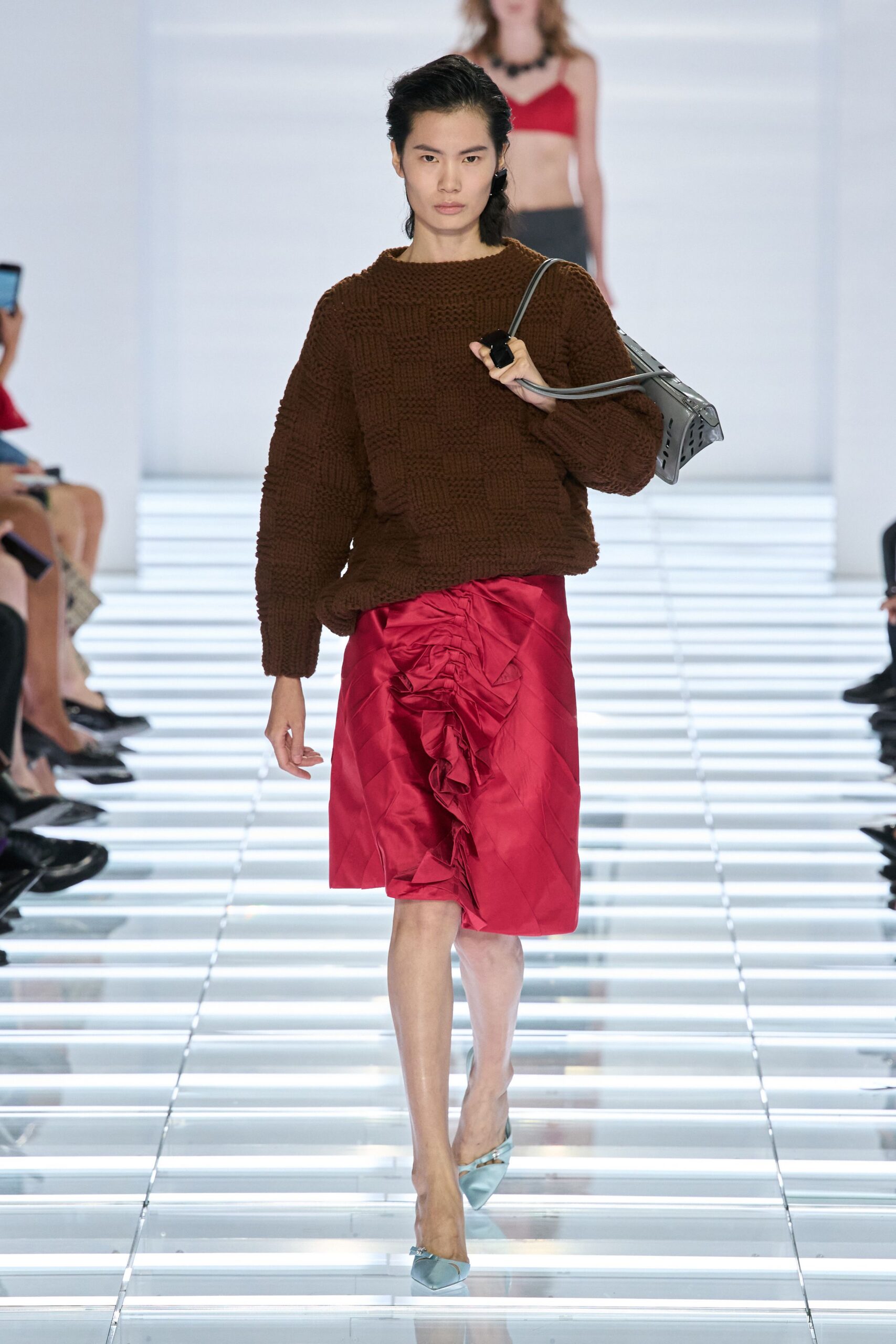 Chocolate chunky knit sweater in a basket-check texture. Slate leather shoulder bag with perforated sides and long tubular straps. Crimson satin knee skirt ruched into a cascading front rosette. Pale blue satin mules with tiny bow hardware. Hotel lobby flowers being changed out after a wedding breakfast.