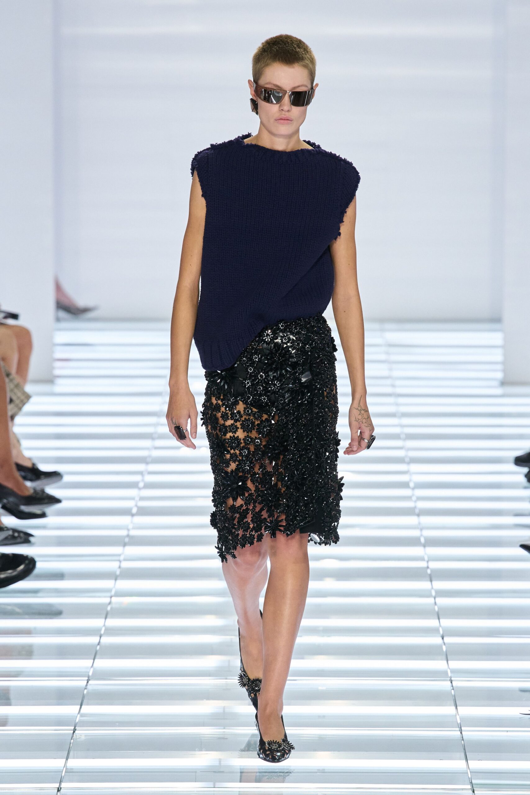 Wraparound black visor lenses with mirrored glare. Ink ribbed knit, cut boxy with rough-fringed cap edges. Black sheer lace skirt crusted with dense floral appliqué and sequins. Pointed pumps topped with black beaded flower bursts. Crossing a hotel atrium before the wedding guests recognize you.