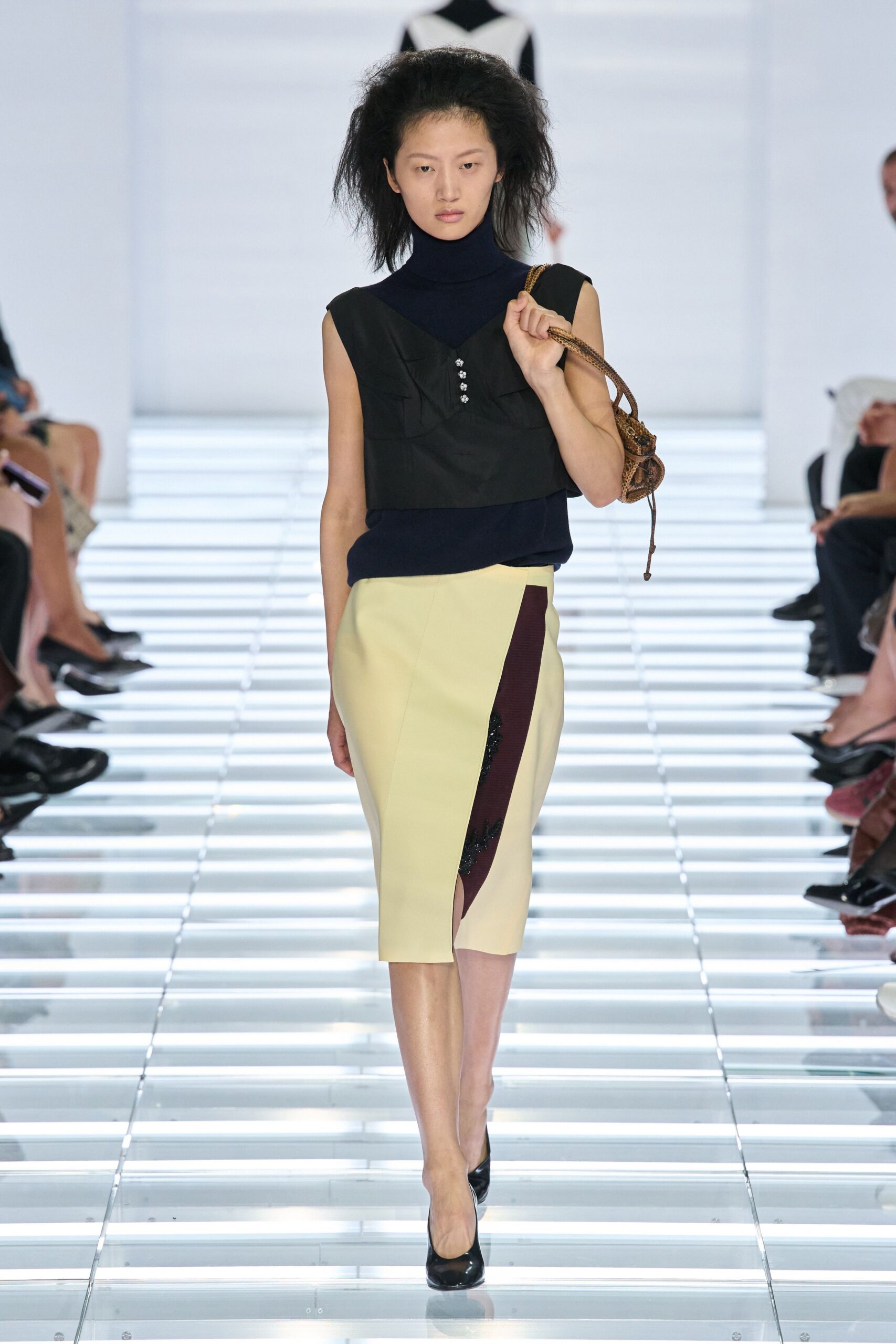 Navy fine-knit turtleneck, close through the neck and armholes. Black cropped bib top with four tiny crystal buttons. Buttercream pencil skirt, wrapped front split by a plum inset. Small brown python bag with a long drawstring tail. Black patent pumps. Museum benefit at noon, still carrying the market bag from earlier.