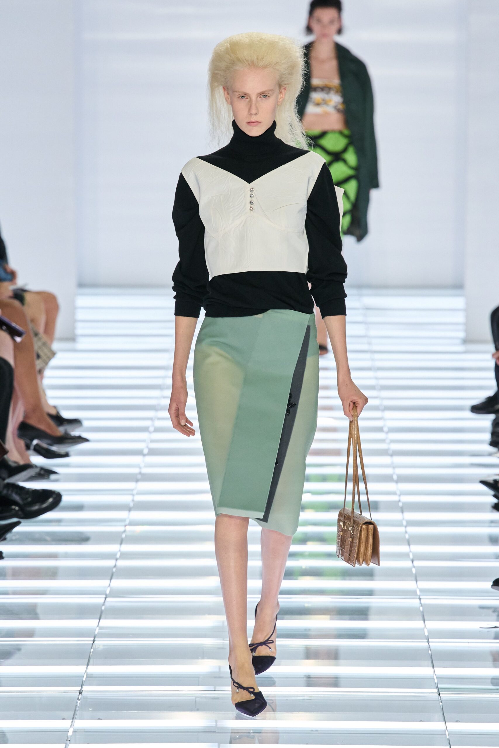 Black knit turtleneck with a white bustier panel, lightly creased. Seafoam pencil skirt cut with a sharp black angled inset. Bronze croc-embossed shoulder bag with long doubled straps. Black pointed flats tied with thin ankle cords. Crossing a glass atrium before the museum opens to the public.