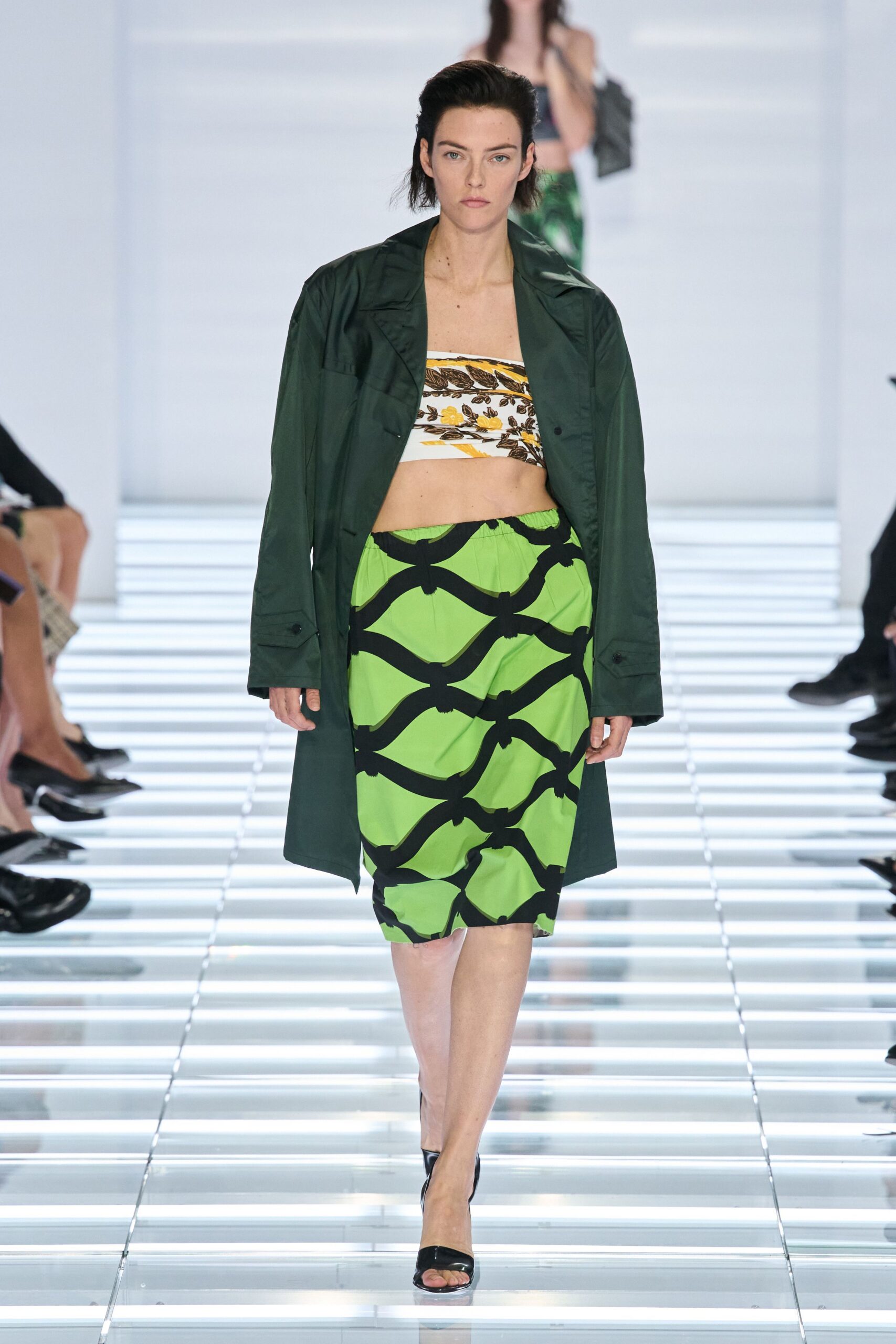 Dark pine nylon coat worn open, flap pockets low at the wrist. White bandeau printed with ochre and brown leaflike bands. Lime pencil skirt crossed with thick black ribbonlike arcs. Black patent peep-toe mules. Walking the glass corridor between pool deck and lobby before dinner.