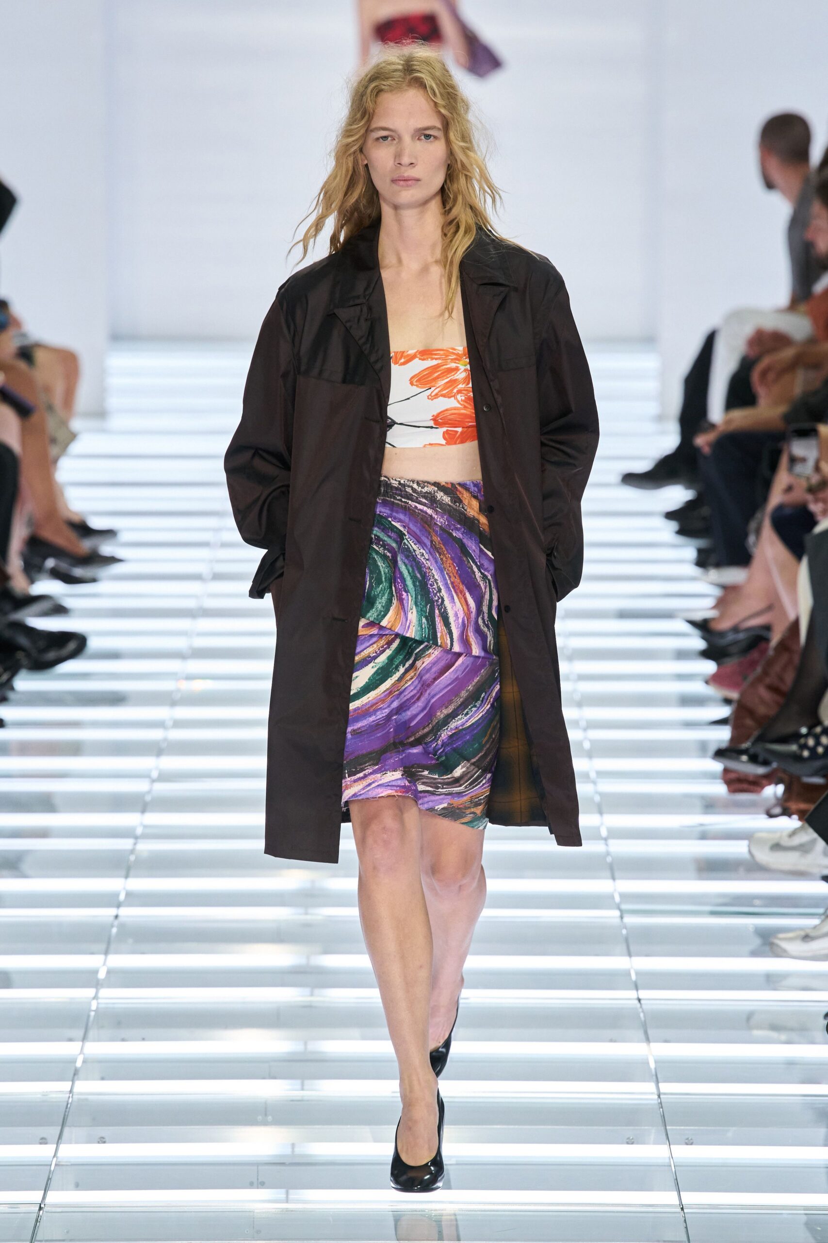 Dark chocolate car coat in crisp technical cloth, patch pockets high on the chest. White bandeau printed with broad orange petals and a fine black stem. Body-skimming mini in violet and jade marbling, hem cutting slightly askew. Black patent pumps. Walking across a museum atrium with rain still drying on your shoulders.