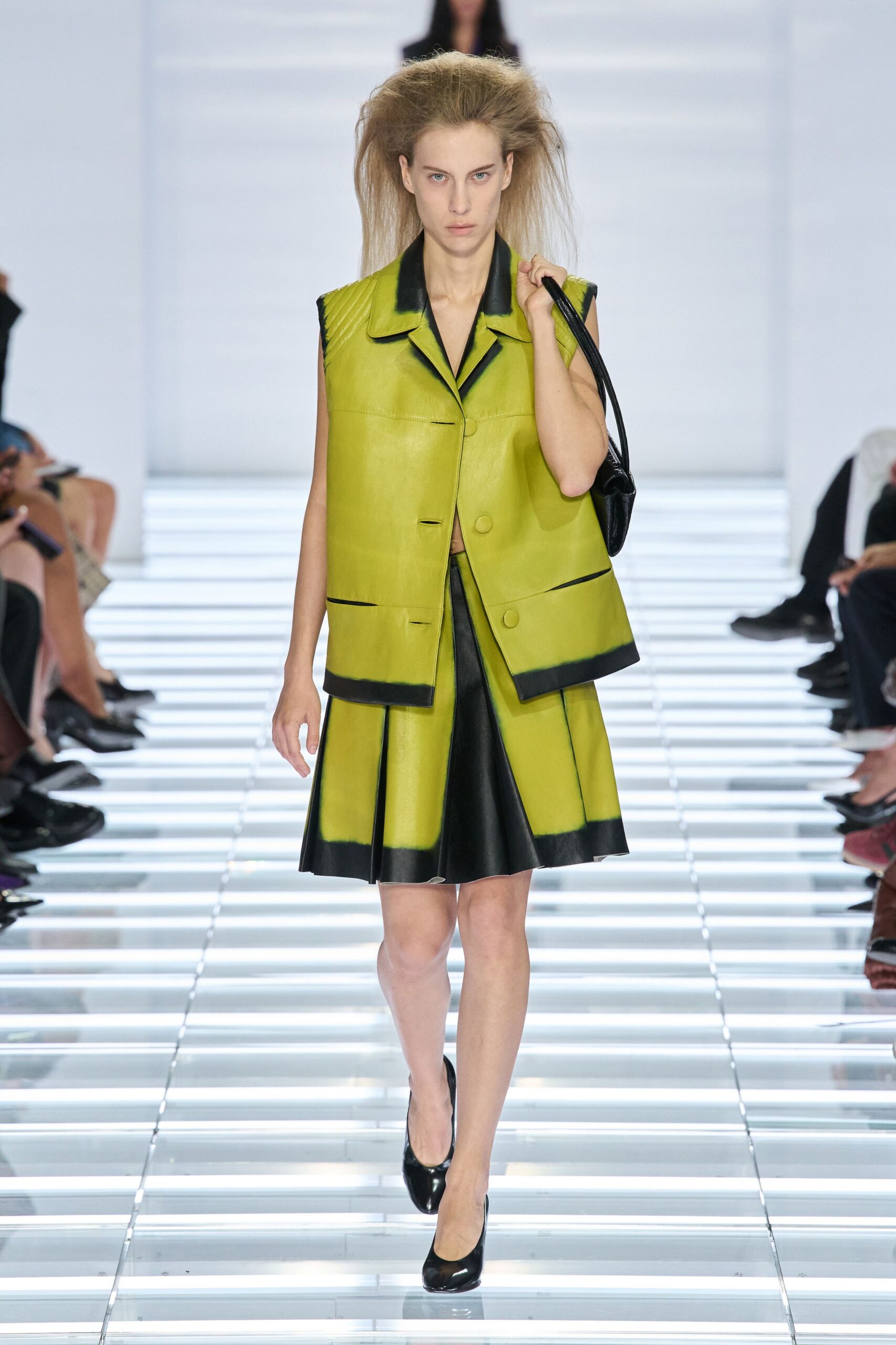 Chartreuse leather vest with quilted shoulders and black dipped edges. Knife-pleated leather skirt, black center panel breaking the acid green. Glossy black shoulder bag tucked tight under the arm. Black patent pumps with a blunt rounded toe. Walking into a municipal lobby still humming with strip lights.