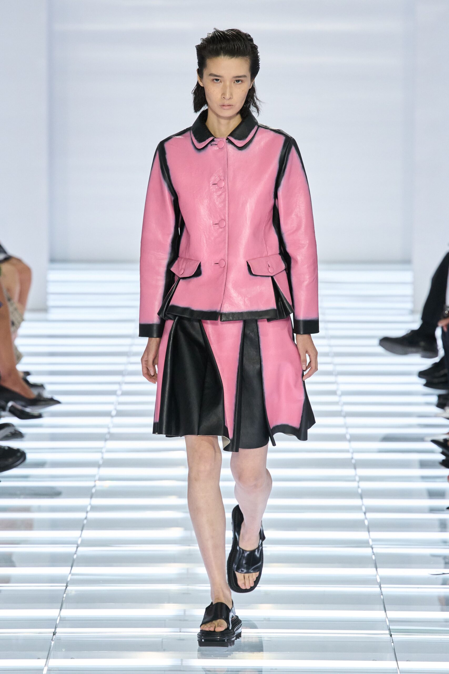 Bubblegum leather jacket with black piping, flap pockets and covered buttons. Matching paneled skirt, black leather godets breaking the pink sweep. Black leather toe-loop sandals on a thick sole. Walking the concourse after the shops shut, floor still throwing back light.