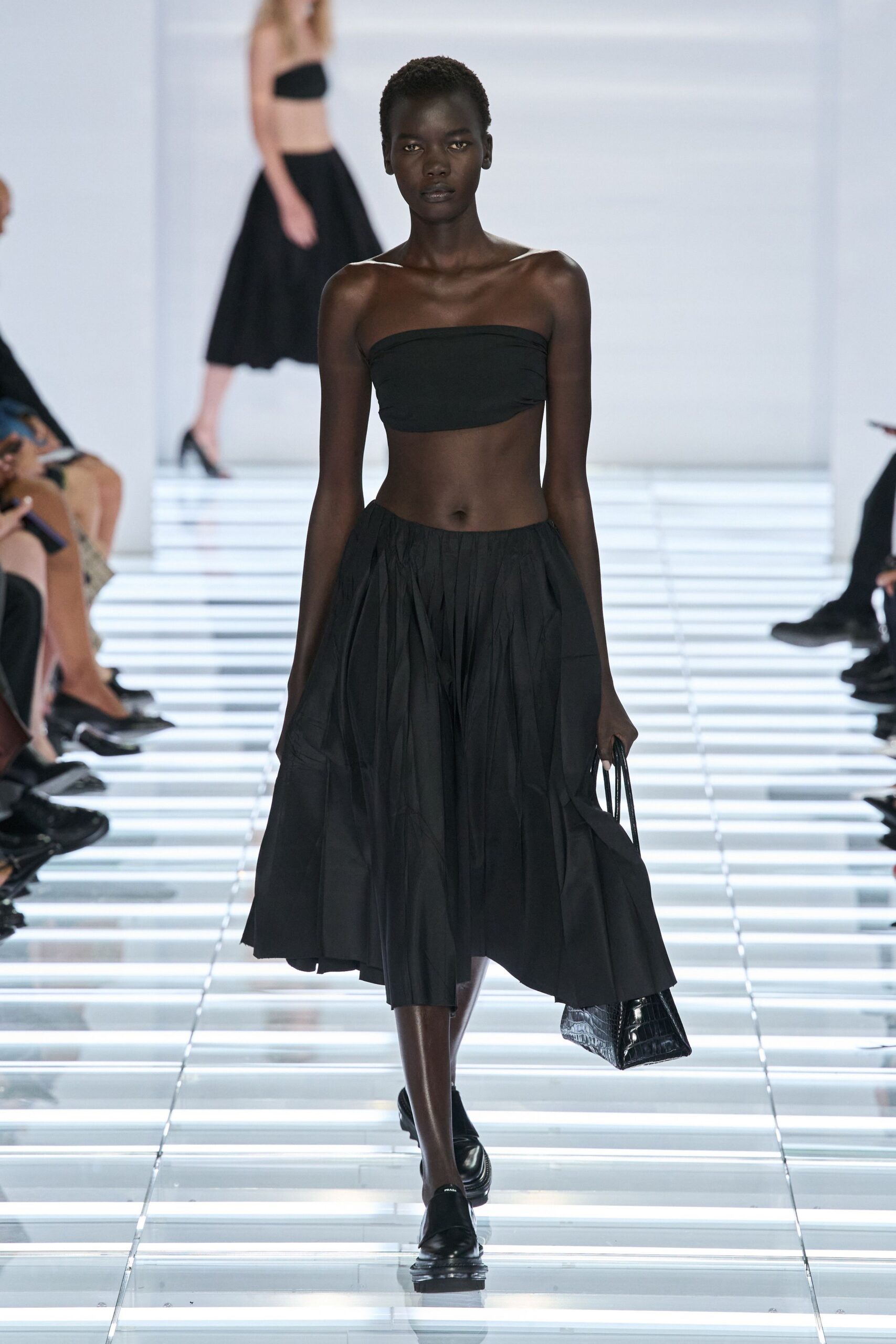 Black jersey bandeau, ruched slightly at one side seam. Voluminous black midi skirt in crisp pleated poplin. Glossy crocodile-embossed clutch hanging from thin straps. Black leather loafers with a thick rounded sole. Walking across a hotel atrium before anyone has started the piano.