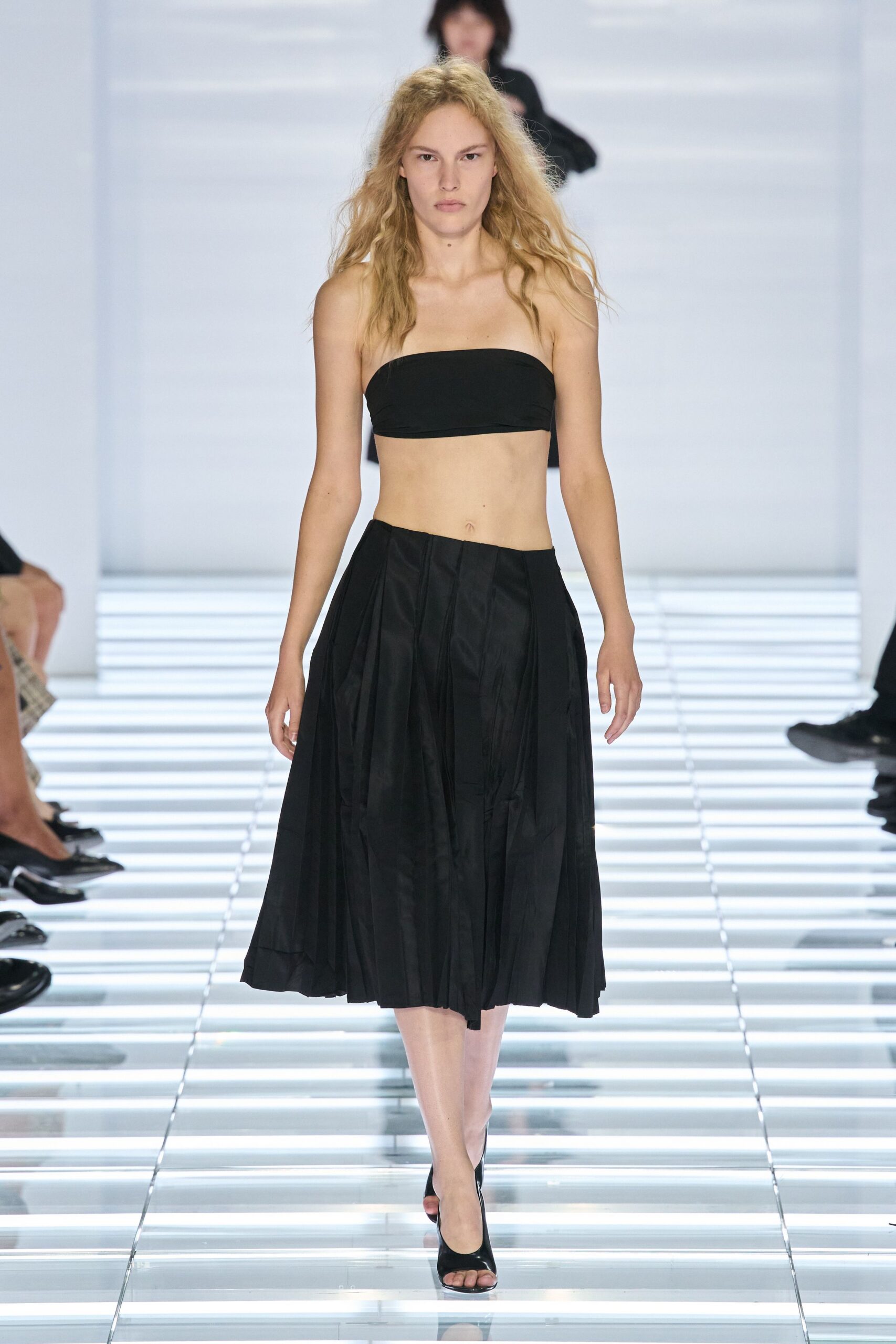 Black jersey bandeau, cut straight and wrapped tight across the bust. Black midi skirt in crisp taffeta, knife pleats falling from a dropped point. Black open-toe mules in glossy patent. Walking the hotel corridor to the ice machine after midnight.