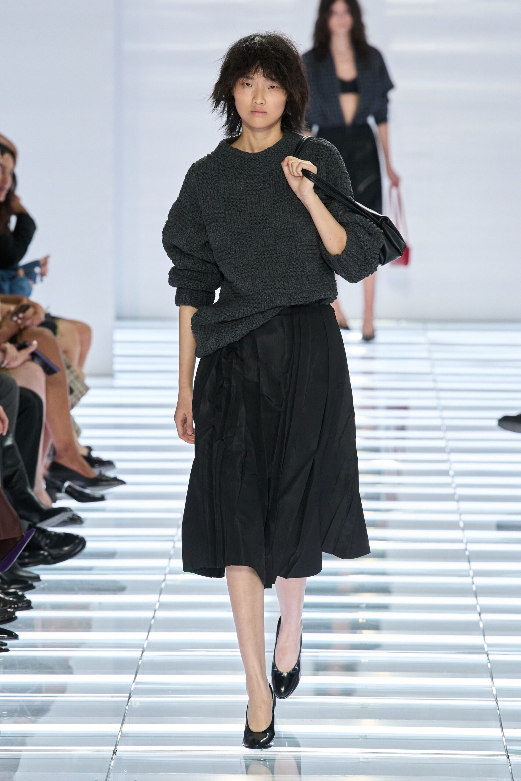 Charcoal open-knit sweater, hem hitched crooked over the waist. Soft black leather tucked tight under one arm. Black midi skirt in crinkled taffeta, hanging in uneven pleats. Black patent pumps. For carrying groceries home after rain, pavement still shining.