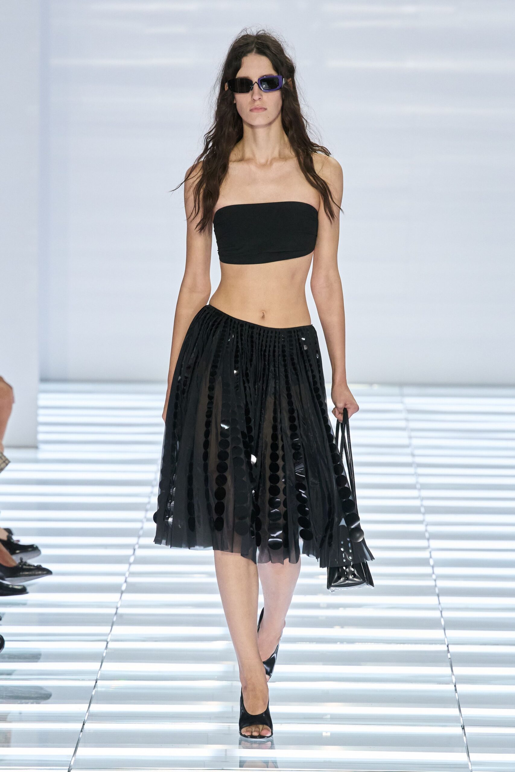 Narrow rectangular sunglasses with one violet outer rim. Black stretch bandeau cut straight across the chest. Sheer black pleated skirt striped with oversized glossy paillettes. Black patent tote with a pierced flap corner. Black peep-toe mules. Walking into a hotel atrium before the conference crowd has finished breakfast.