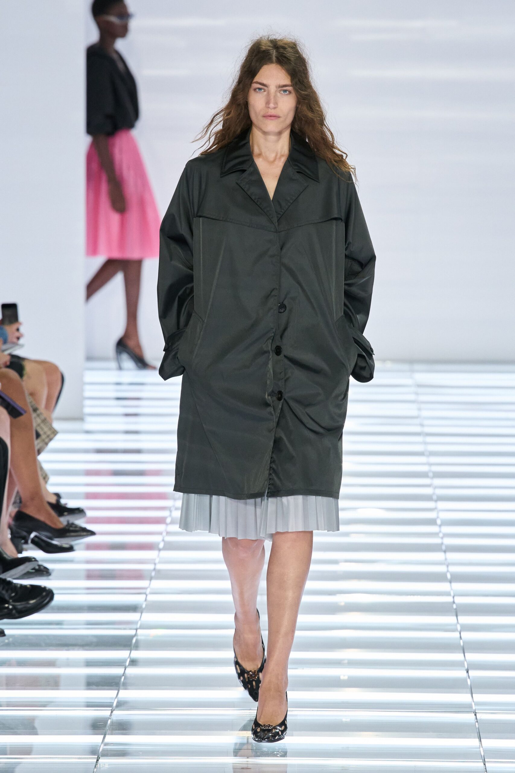 Charcoal technical coat with a satiny collar and skewed black buttons. Pale grey pleated hem peeking below the coat. Black cutout pumps with a jeweled vamp clasp. Walking across a hotel atrium before the florist has finished the lobby.