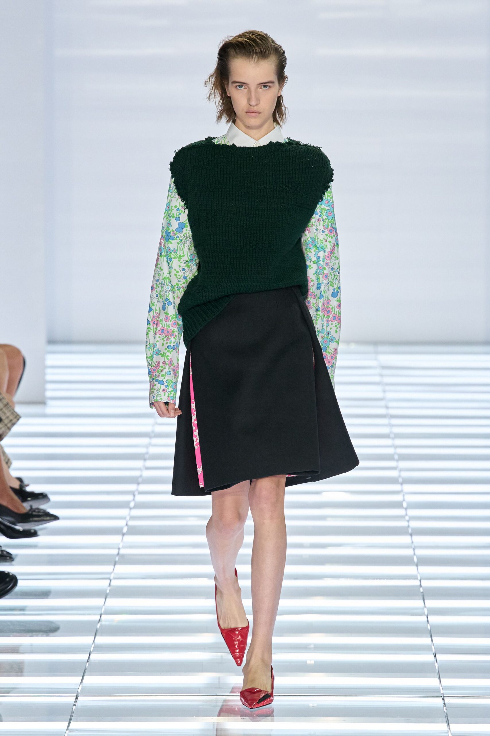 White cotton shirt printed with mint, blue and pink meadow flowers. Dark bottle-green chunky knit, shoulder edges left slightly nubby. Black wrap skirt opening to a hot-pink floral underpanel. Red patent pointed pumps with lightly creased vamps. Crossing the conservatory café before the rain lets up.