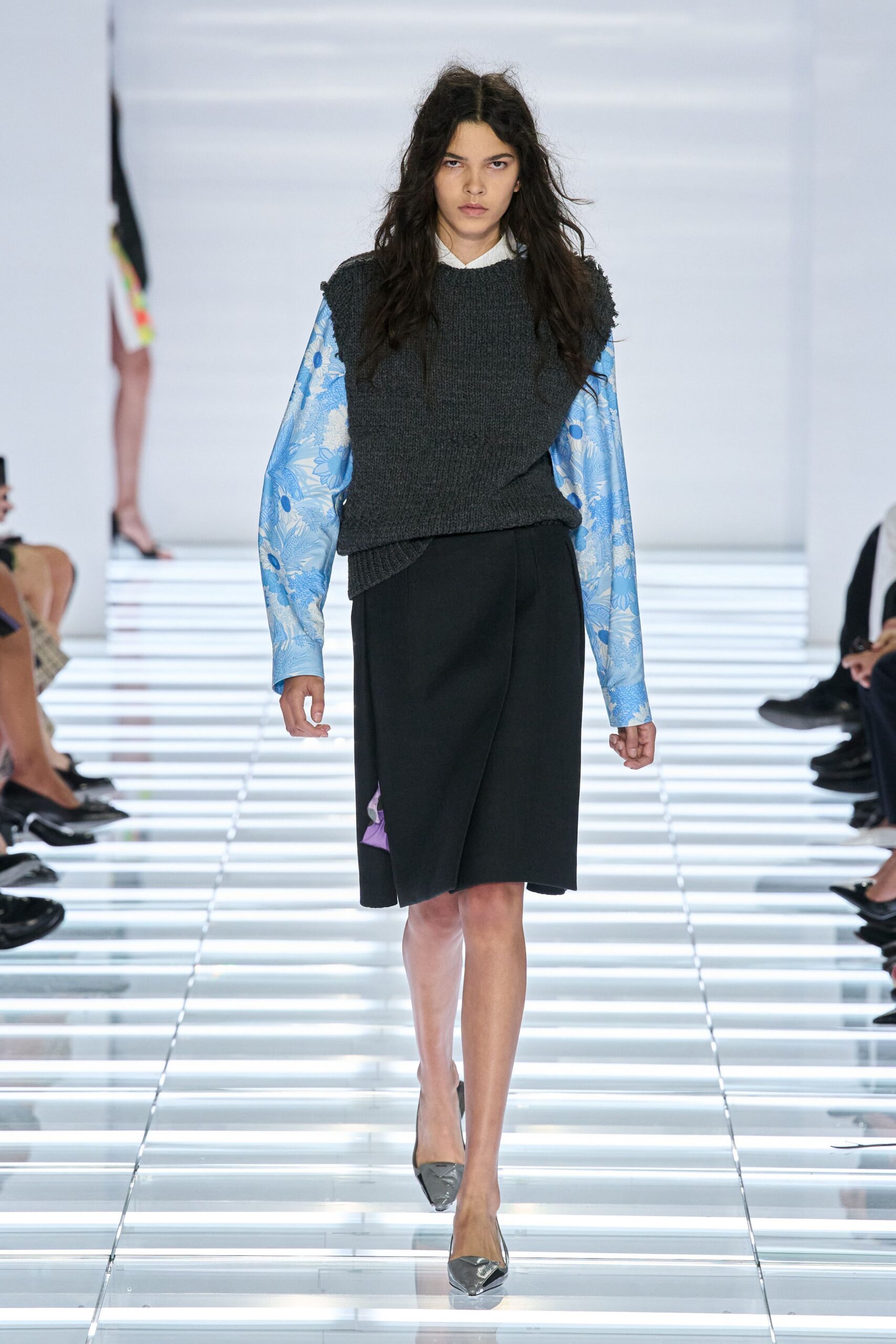 Blue floral jacquard shirt, white collar peeking clean above the knit. Charcoal ribbed sweater vest, one side drooping into an uneven wrap. Black wrap skirt with a lilac lining flash at the side slit. Silver pointed pumps. Office atrium at lunch, carrying spring under a winter layer.