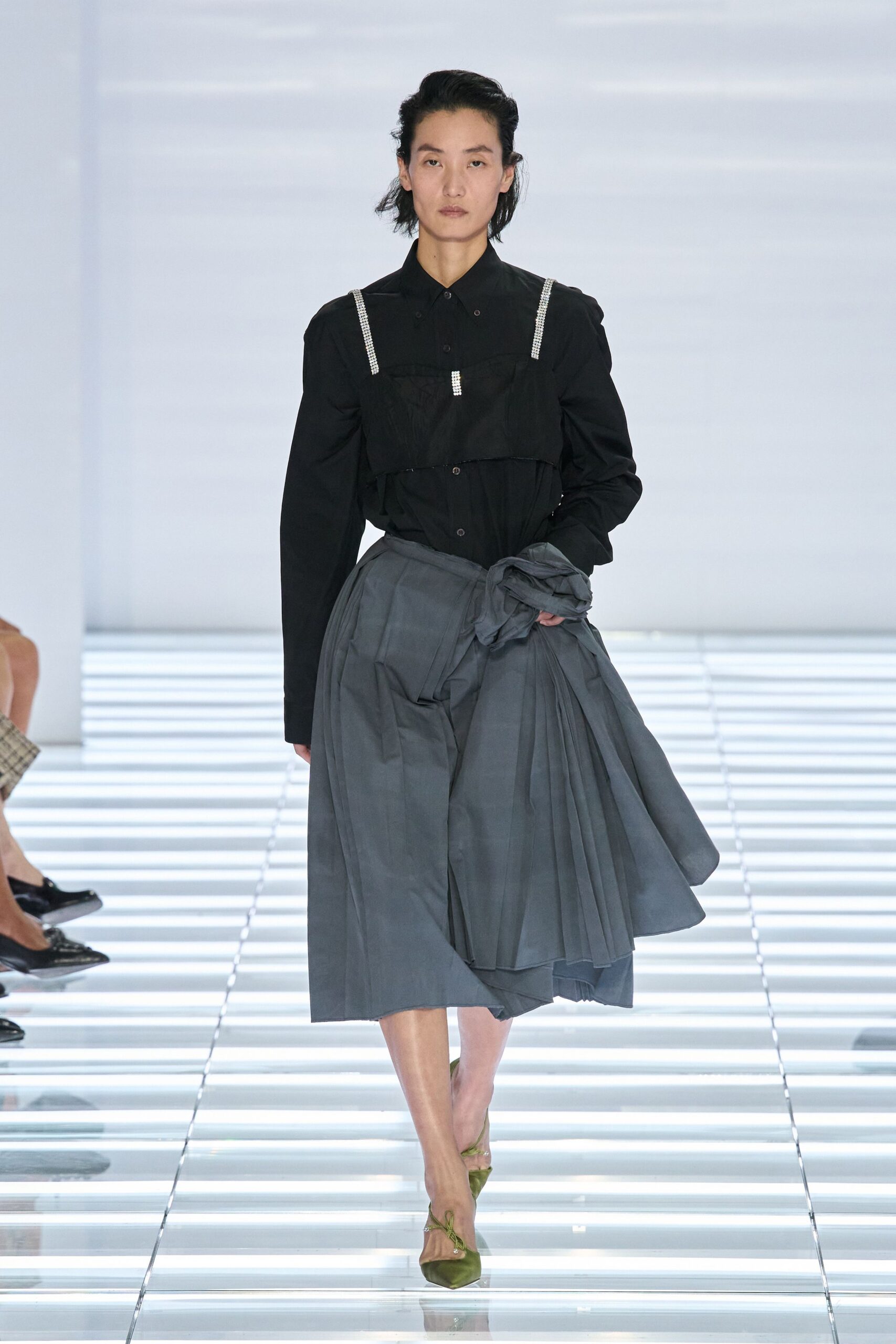 Black cotton button-up with a bib overlay and rhinestone shoulder straps. Slate wrap skirt, knife pleats breaking from a twisted side knot. Moss satin pumps with thin ties crossing the instep. Crossing a glass atrium before the office air has warmed up.