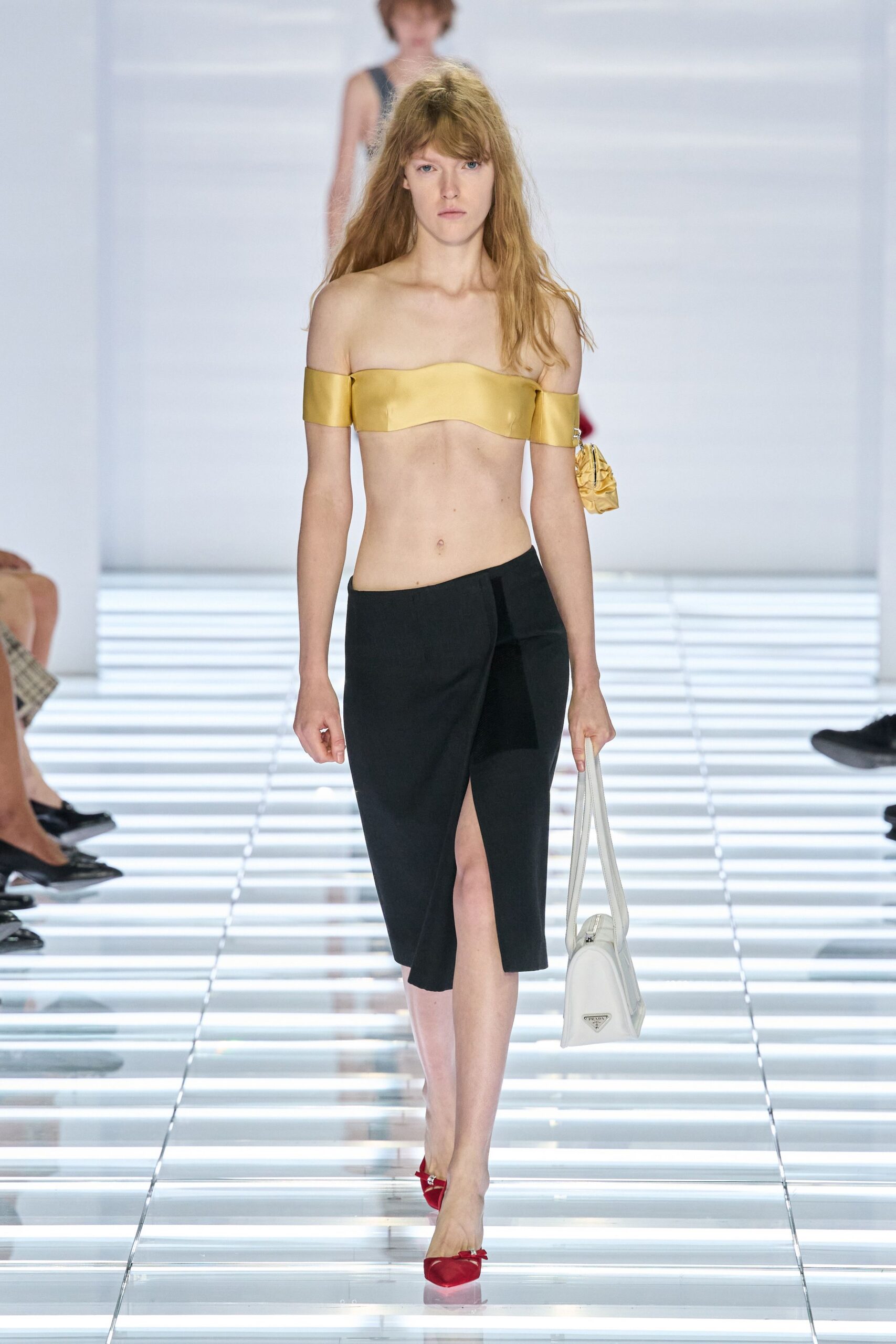 Butter-yellow satin bandeau with off-shoulder bands clipped at one sleeve. Black wrap pencil skirt, front split opening from a low overlap. White leather bowling bag with long doubled straps. Red satin pumps with a tiny silver buckle. Walking into a hotel lobby still warm from the pavement outside.