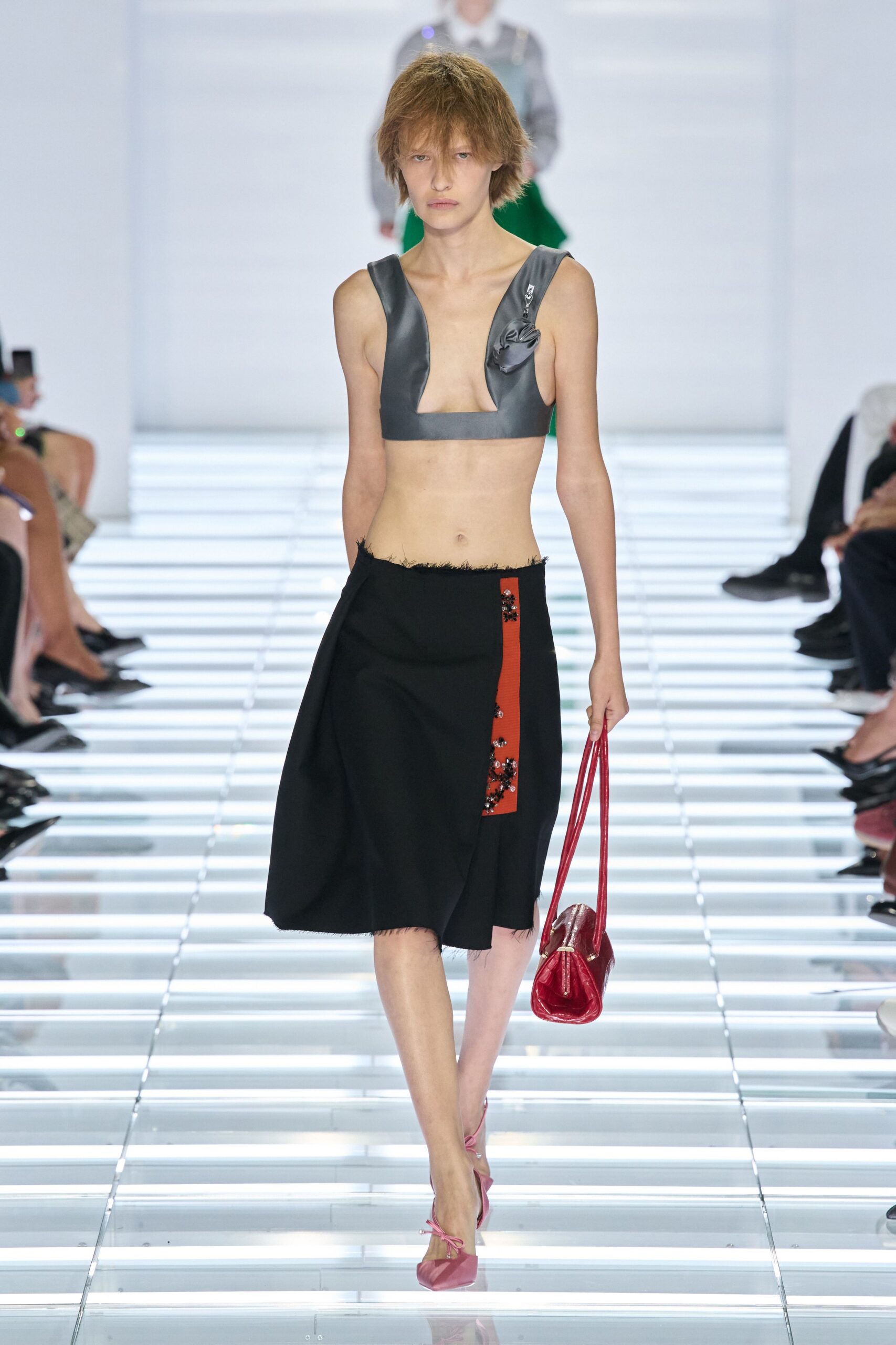 Slate satin crop top, plunge cutouts and a pinned rosette by the strap. Black wrap skirt with frayed edges and a rust tab beaded in clusters. Glossy red patent barrel bag on narrow shoulder straps. Pink satin pumps tied with thin laces at the ankle. Crossing a glass footbridge after rain with the wrong bag for groceries.