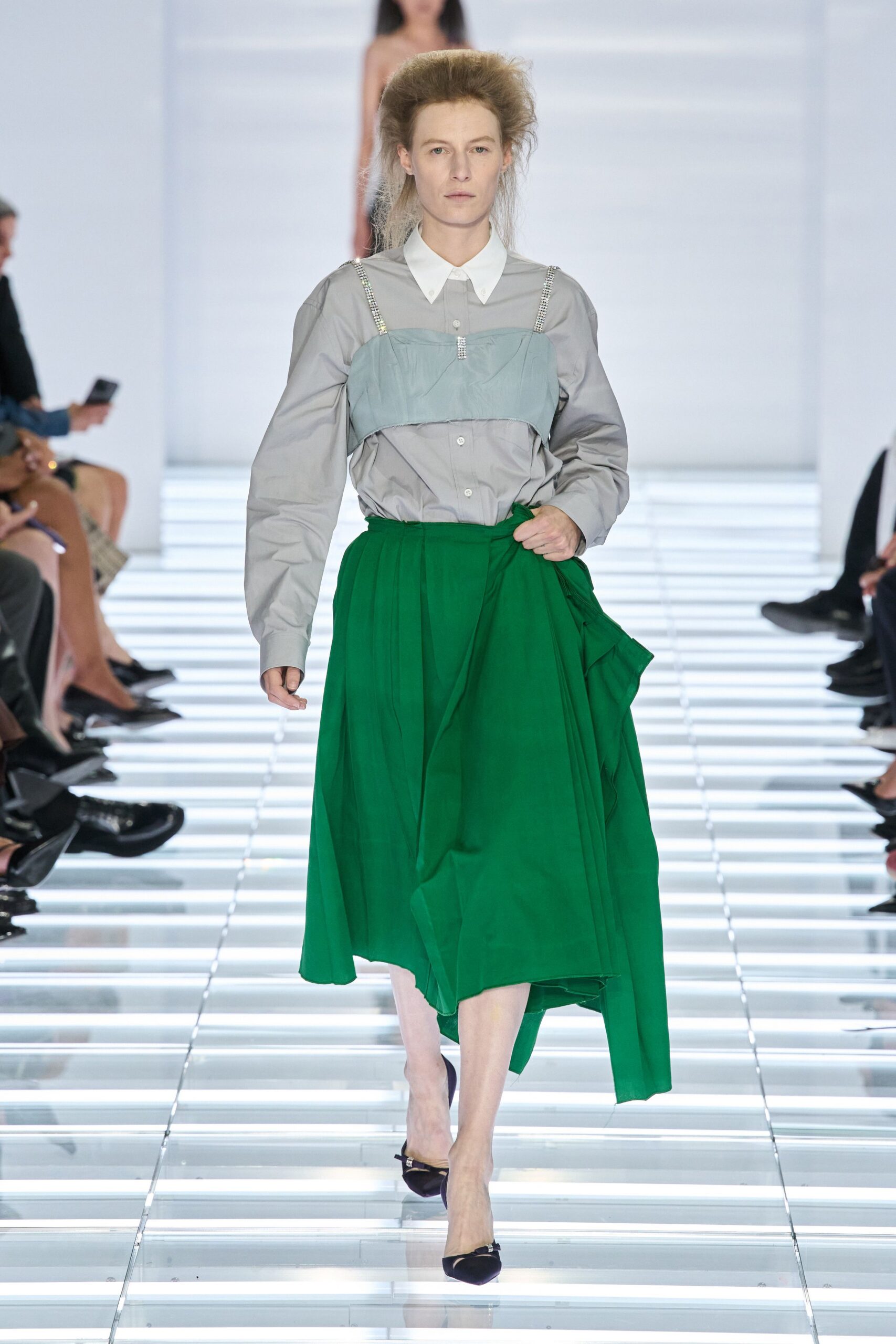 Pale grey poplin shirt with a sharp white point collar. Dusty blue bandeau overlay hung from rhinestone shoulder straps. Emerald wrap skirt in fluid crepe, one panel trailing loose. Black satin pumps with a tiny crystal buckle. Crossing a hotel atrium after noon, still carrying yesterday's room key.