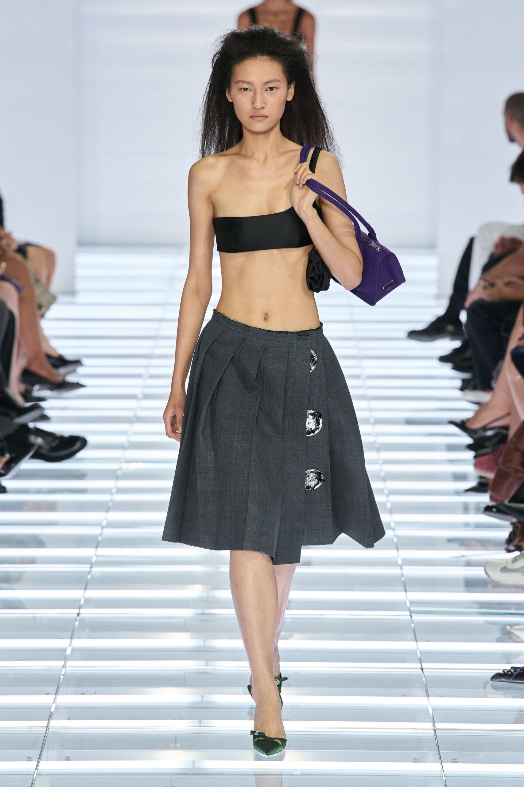 Black bandeau cut dead straight across the chest. Charcoal wool skirt with deep inverted pleats and raw waistband edge. Purple nylon shoulder bag with a small silver corner plaque. Dark green satin pumps with tiny bow trims. Walking into a department store cosmetics hall before it opens.