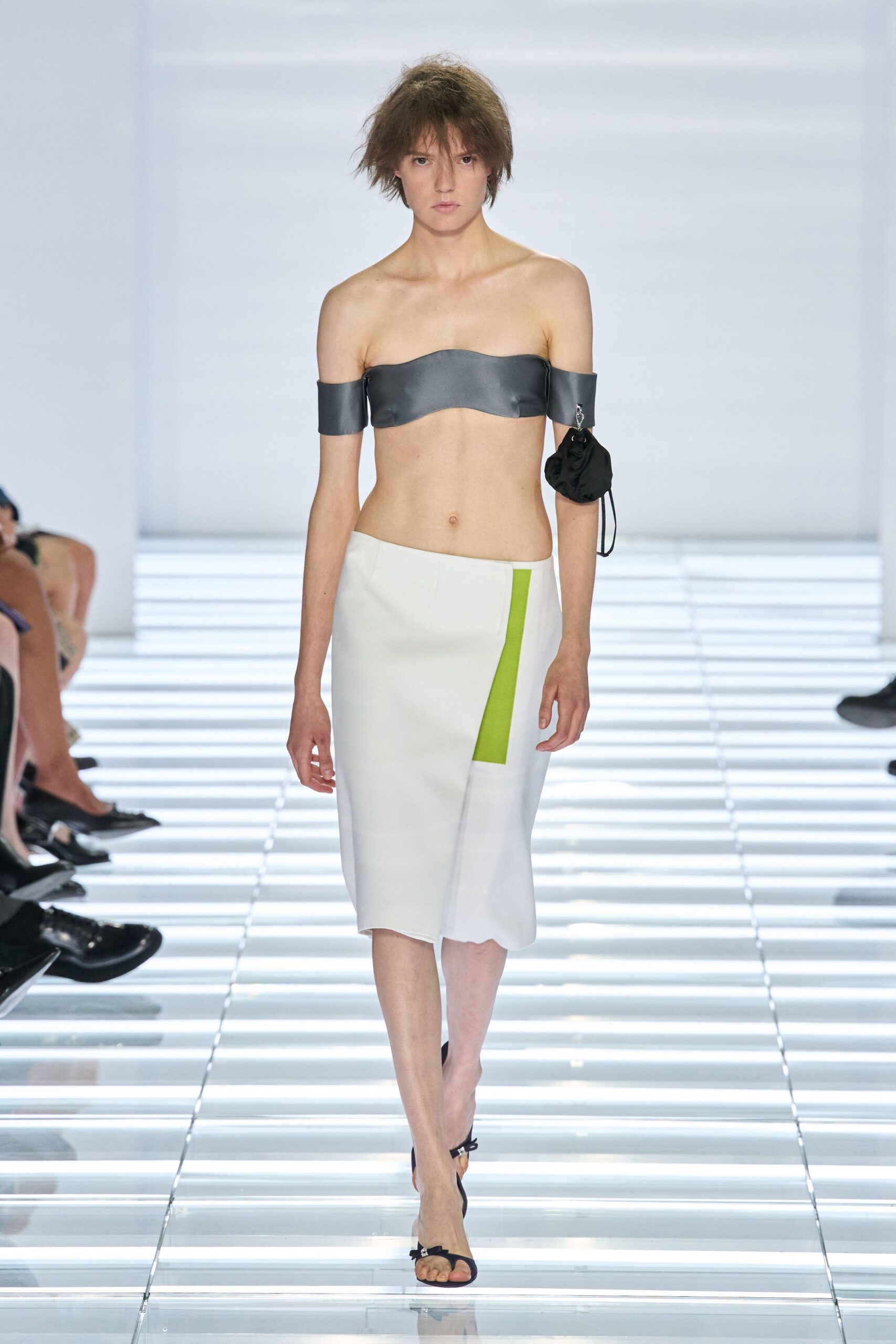 Steel-grey satin bandeau with off-shoulder bands wrapping both upper arms. Black drawstring pouch clipped beneath the right sleeve band. White midi skirt cut on a slant, lime inset flashing at the hip. Black thong sandals with a tiny bow at the toe. Crossing a glass corridor before the office plants get watered.