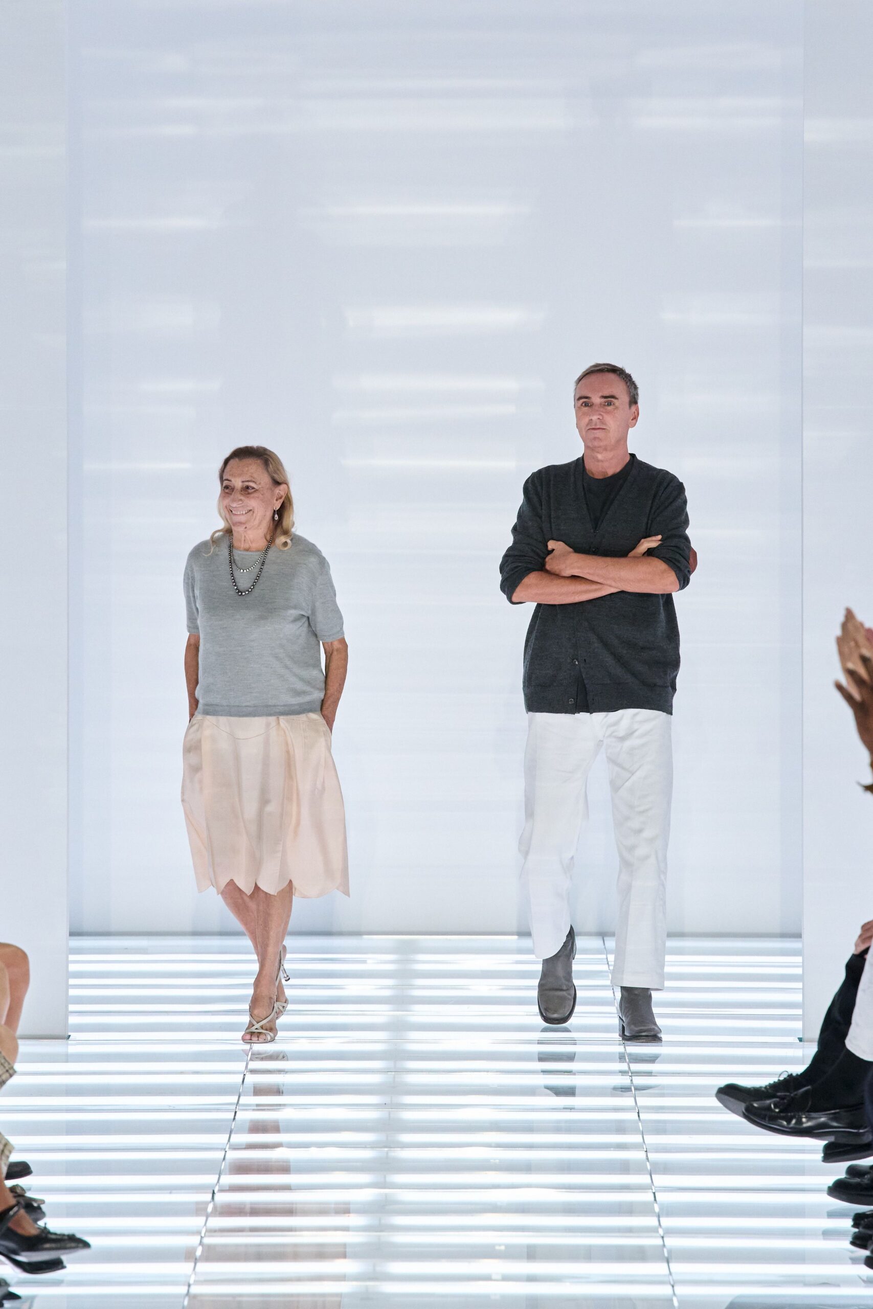Pale grey fine-gauge short-sleeve knit with a clean ribbed hem. Long dark beads doubled once at the collarbone. Blush knee-length skirt with a softly scalloped bubble hem. Narrow white slingback sandals with slim crossed straps. Crossing a glass corridor before the museum opens to the public.