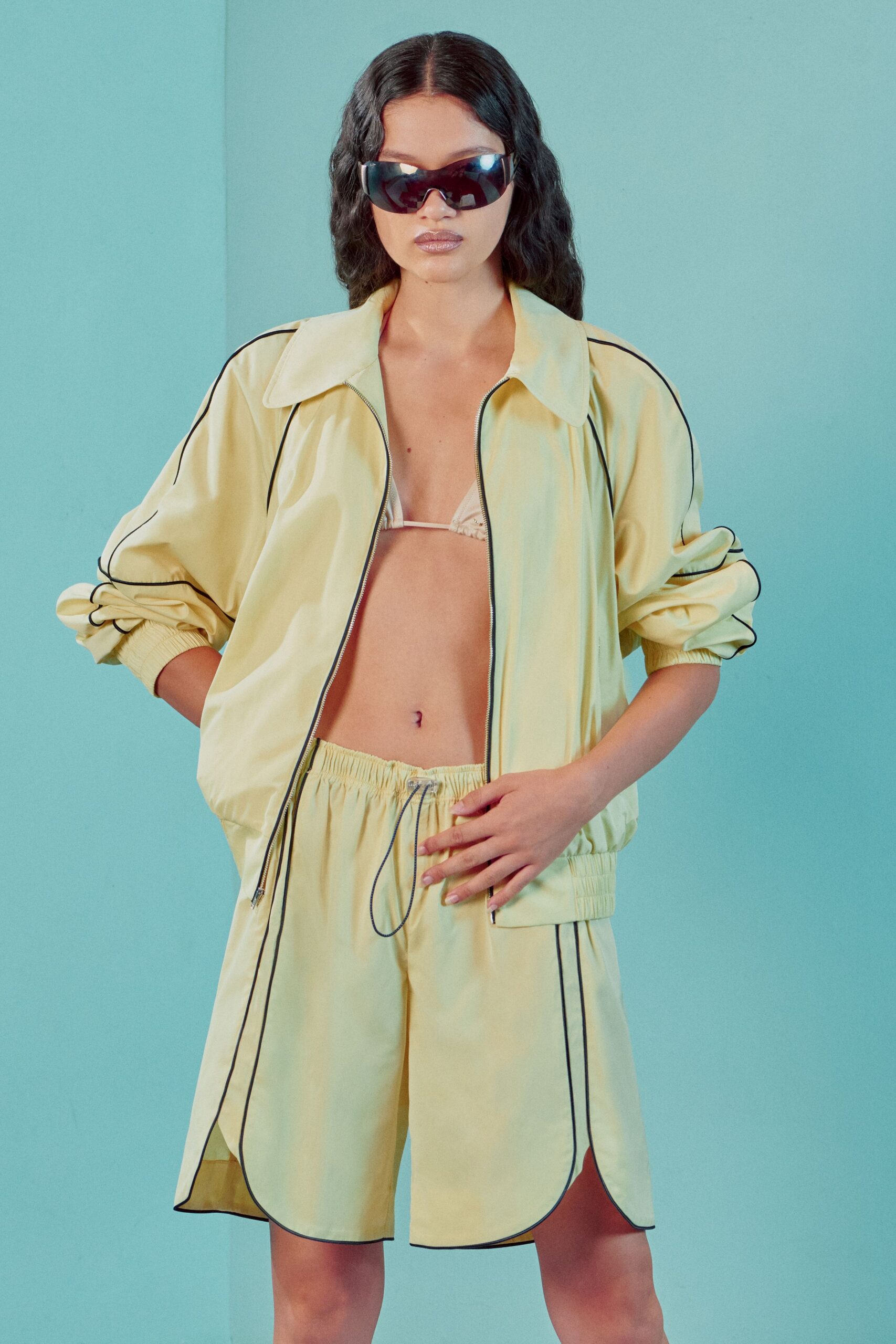 Wraparound black visor lenses. Buttercream satin bomber, black piping tracing the dropped sleeves. Tiny ruched triangle top peeking through the open zip. Matching satin track shorts with a long cord drawstring. Poolside corridor, noon heat bouncing off painted concrete.