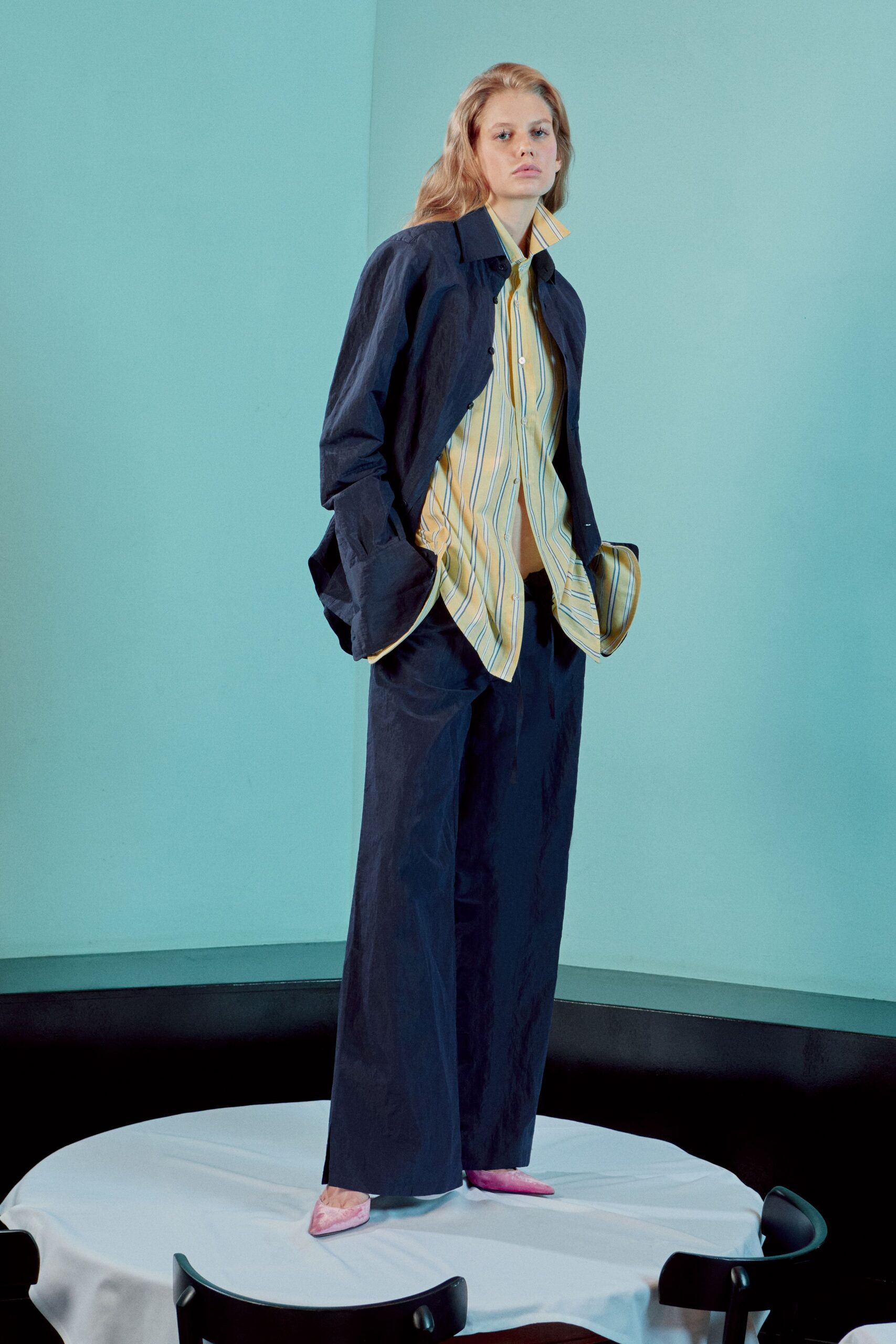 Butter-yellow striped shirt, collar kicked up and hem hanging open. Ink poplin jacket cut short, sleeves pooling over the hands. Matching ink trousers in crisp cloth, falling straight from the hip. Pink velvet pointed flats. Standing on a restaurant table before the lunch crowd is let in.