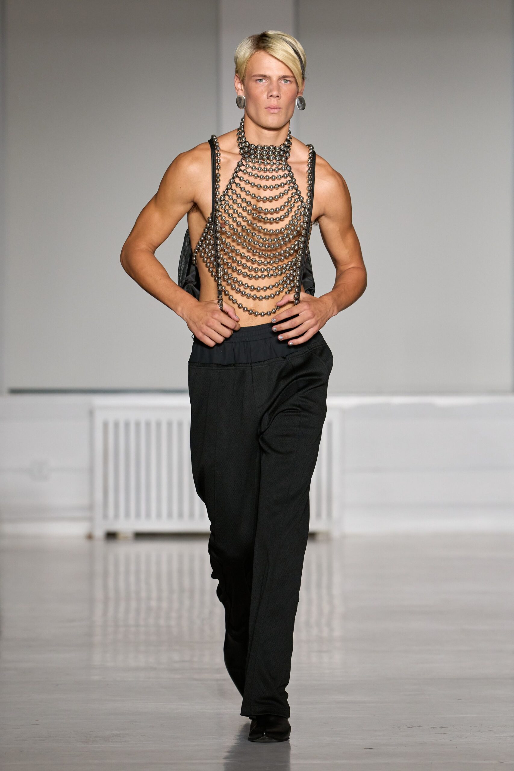 Gunmetal disc earrings, flat and oversized against the jaw. Gunmetal ball-chain bib cascading in dense scallops to the waist. Black straight trousers with a soft gathered waistband slung low. Black pointed boots, tips just breaking under the hem. Walking into a basement party after midnight with nowhere to put your shirt.