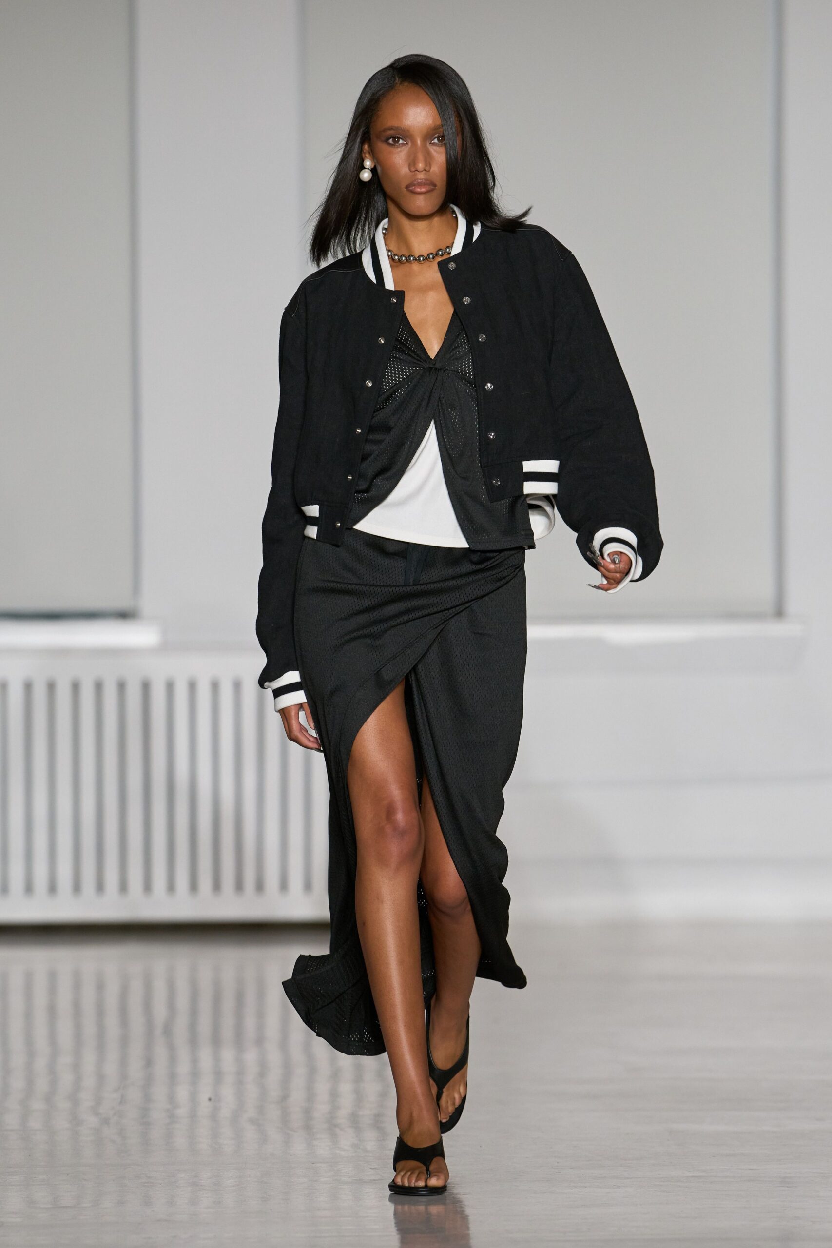 Black bomber with white striped rib cuffs and silver snap front. Black mesh top knotted at the bust over a white inset. Black athletic-mesh skirt, draped into a high thigh slit. Short gunmetal bead strand. Oversized double pearls. Black thong sandals. Walking across a hotel lobby before anyone else is properly awake.