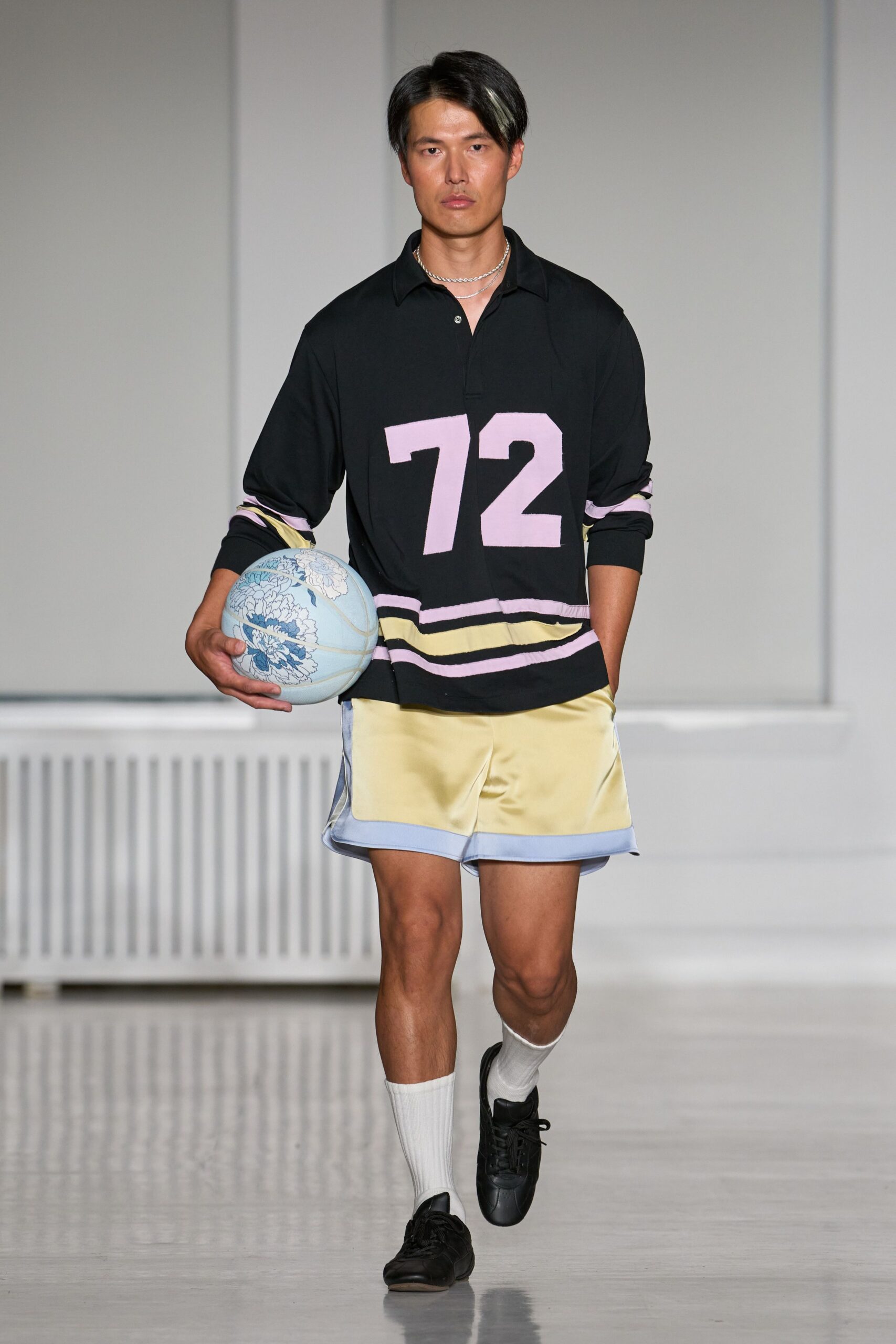 Black rugby shirt with a pale pink 72 and striped hem. Double silver chain at the collarbone. Butter-yellow satin shorts with powder-blue bound hems. White ribbed crew socks. Black leather lace-up trainers. Powder-blue basketball printed with blue peonies. Walking to the park court with florist-wrap still on the bouquet.