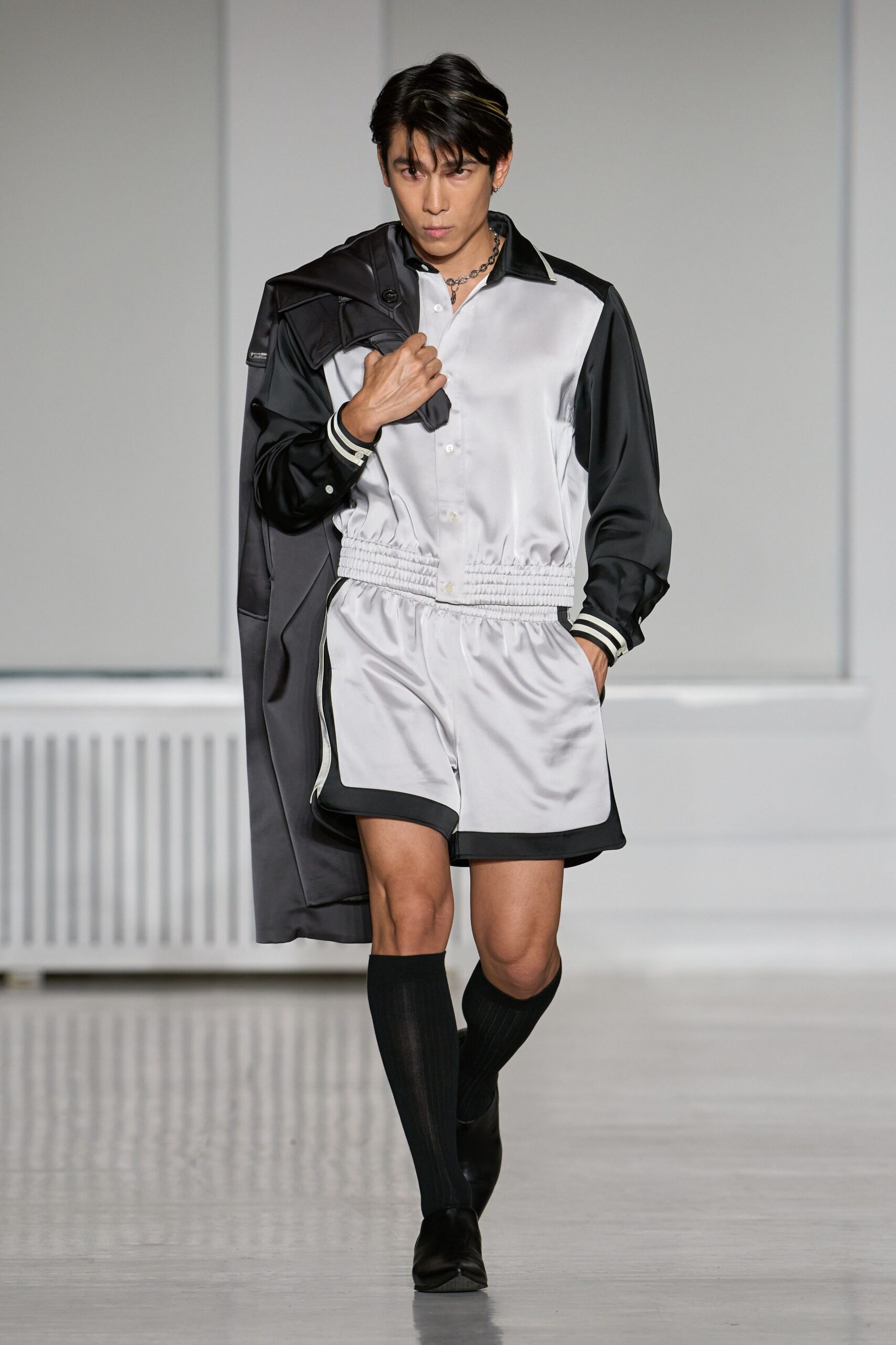 Charcoal satin coat slung over one shoulder, zip tab catching light. Pearl satin romper with black sleeves and a shirred drop waist. Chunky silver chain at the open collar. Black ribbed knee socks. Square-toe black loafers. For taking the hotel stairs when the lift is out again.