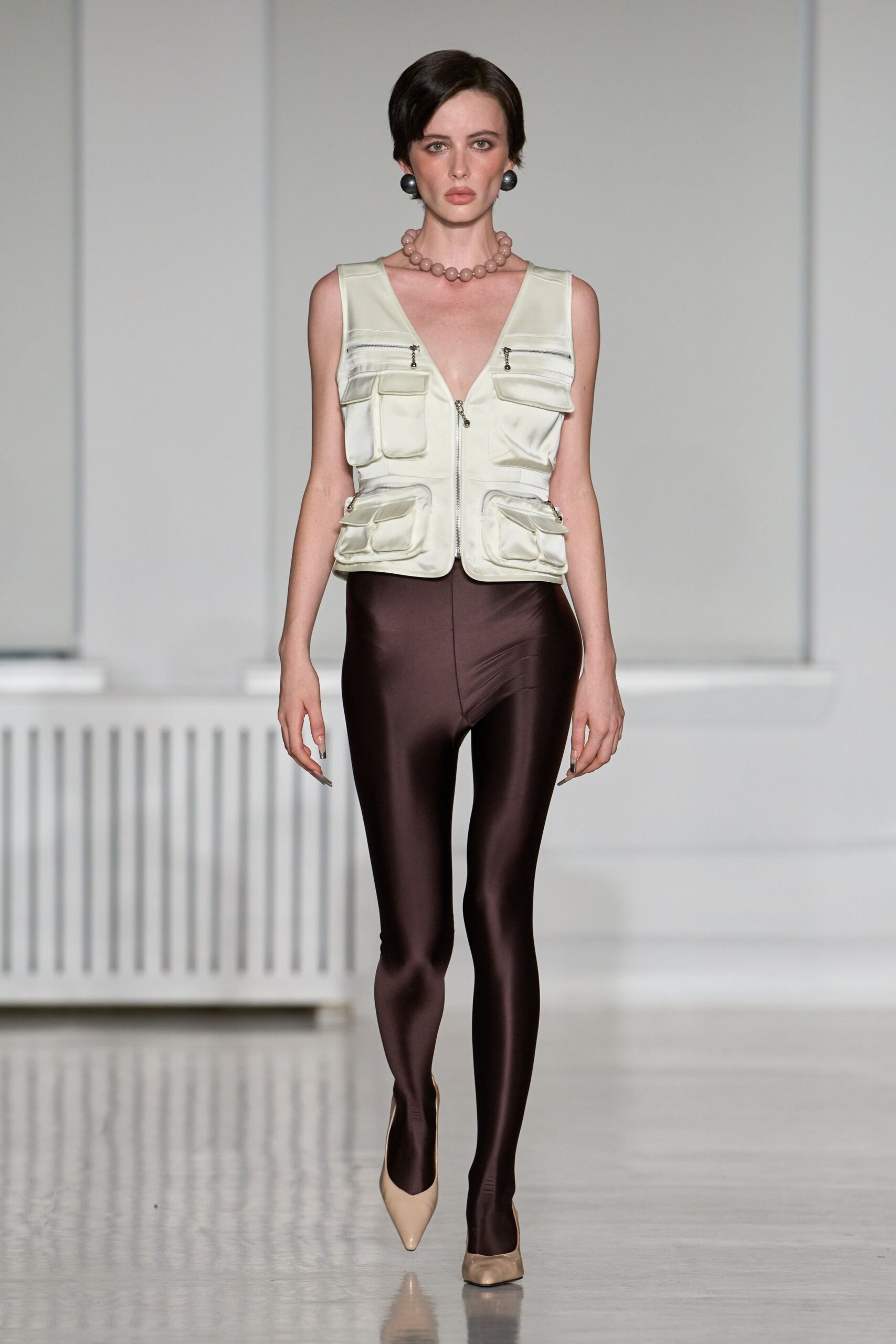 Oversized charcoal ball studs. Powder-pink beads sitting close at the collarbone. Ivory satin utility vest, zipped front and layered bellows pockets. Espresso stretch leggings with a wet gleam and stirrup-tight pull. Nude pointed pumps. For crossing a hotel lobby at dawn with room service trays still outside doors.