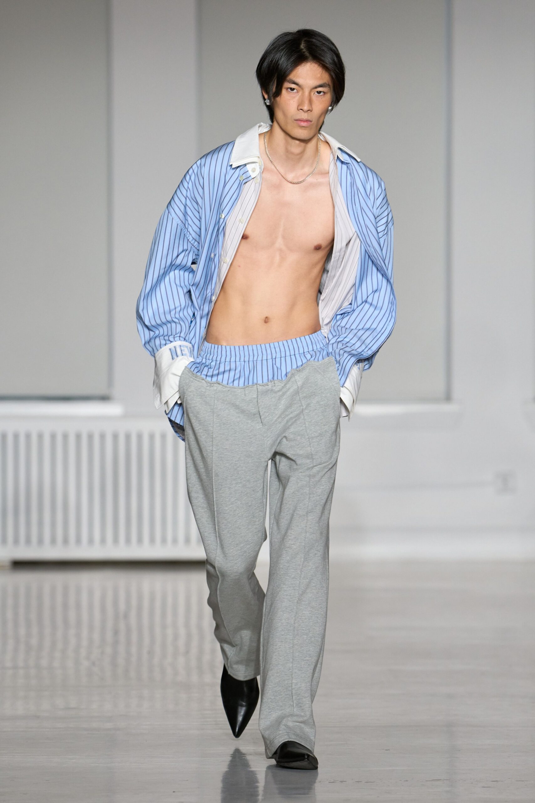 Sky-blue striped poplin shirt worn open, white collar and deep cuffs slipping wide. Heather-grey jersey trousers with a blue striped waistband peeking above. Black pointed boots under the pooling hem. Fine silver chain at the collarbone. For the first coffee run after sleeping at somebody else's apartment.