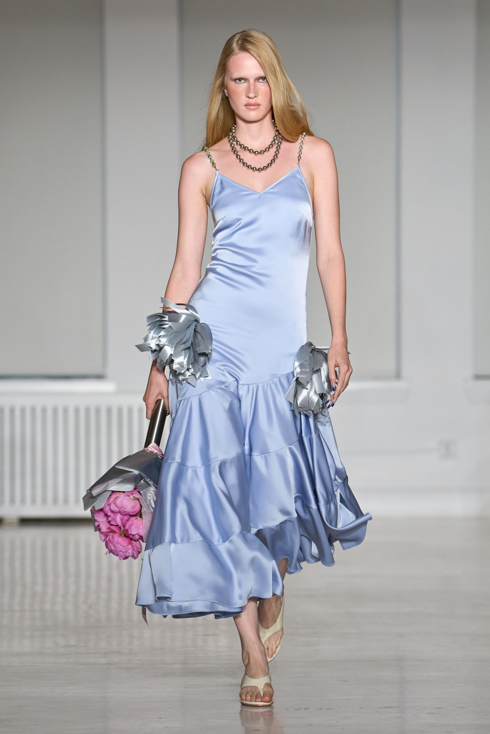 Double strand of gunmetal beads at the collarbone. Powder-blue satin slip dress, chain straps and a low ruffled flounce. Silver ribbon pompons tied at both wrists. Pink peonies wrapped in crinkled silver paper. Bone thong sandals. Leaving city hall with the flowers still in their paper.