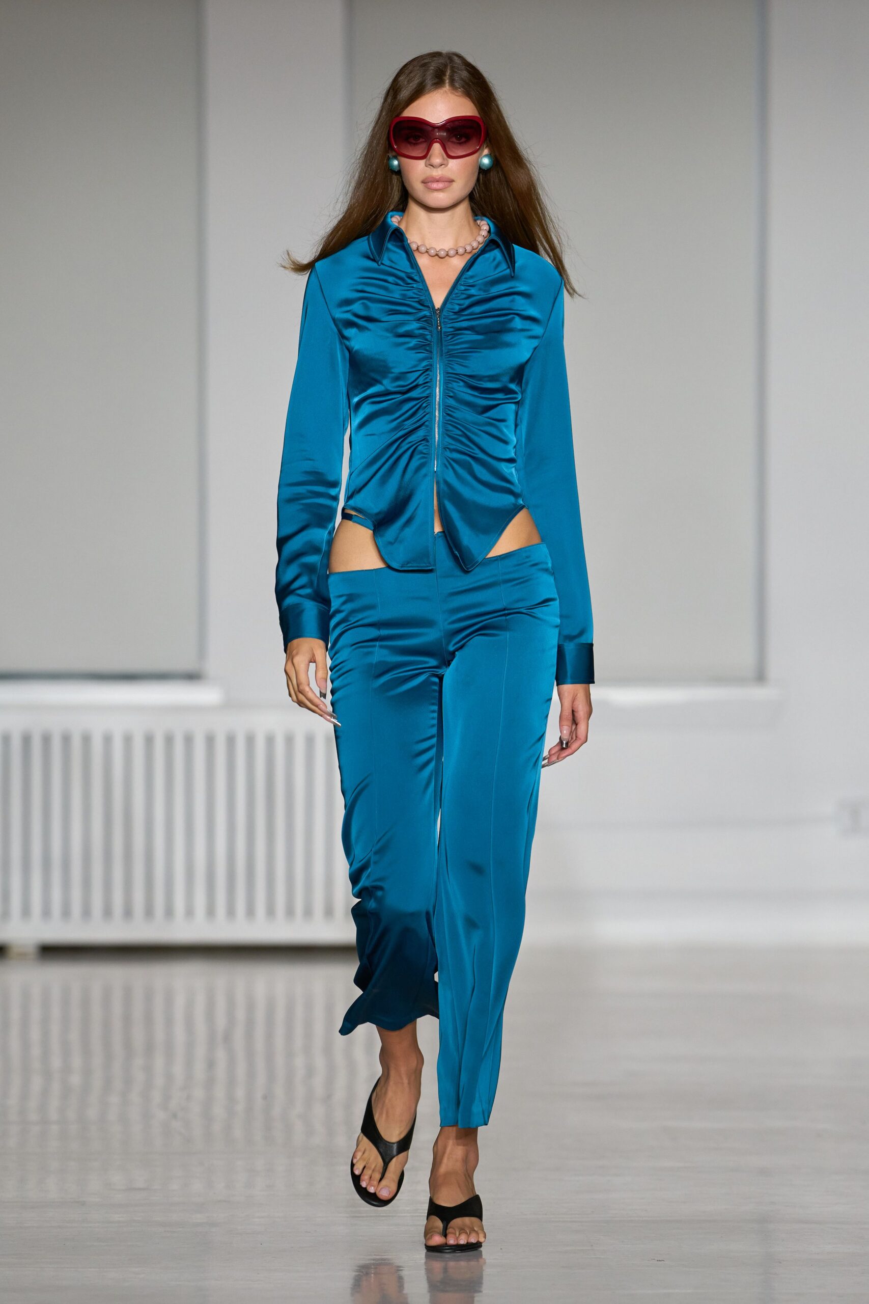 Oversized wine-tinted shield sunglasses with a soft hearted curve. Peacock satin zip shirt, ruched tight along the center front. Matching low-rise satin trousers with sharp hip cutouts. Turquoise ball studs. Short string of round pearls. Black thong heels. Walking into a hotel atrium before anyone else is down for breakfast.