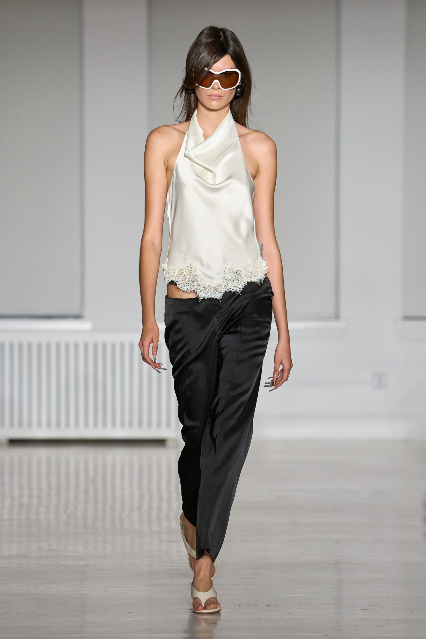 White wraparound frames with amber shield lenses. Oversized black orb drops. Ivory satin halter with a folded cowl and eyelash-lace hem. Black satin trousers, cut low with a twisted overlap at the waist. Cream thong mules. Checking into a seaside hotel before the room is ready.