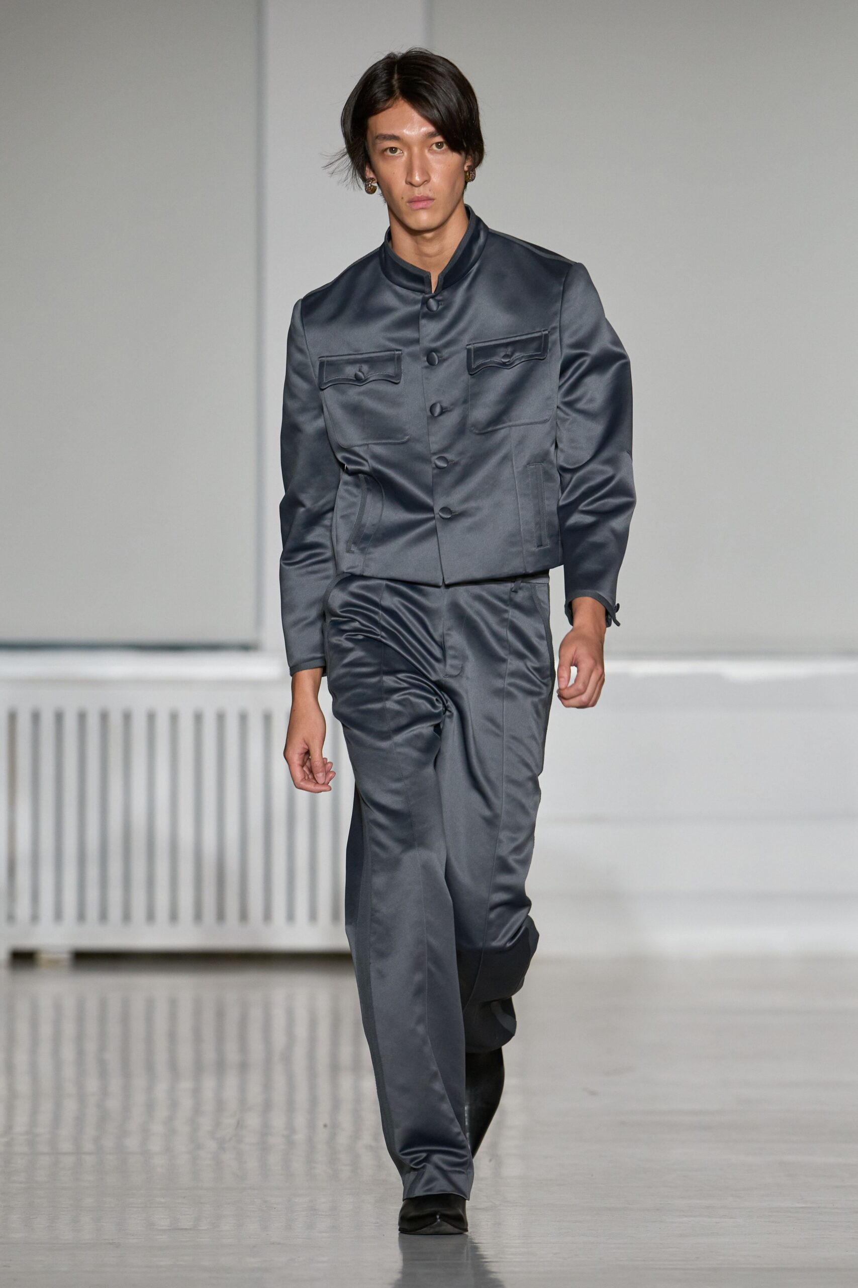 Steel-grey satin jacket with stand collar and covered buttons. Matching satin trousers, pressed crease breaking into soft thigh ripples. Black square-toe boots. Small bronze drop earrings. Hotel corridor at dawn, still wearing last night's formal idea.